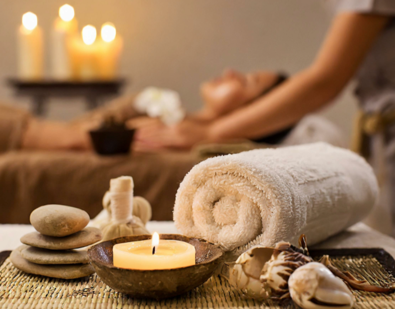 Spa scene with rolled towel, lit candle, stones, seashells, and a woman receiving a massage in the background.