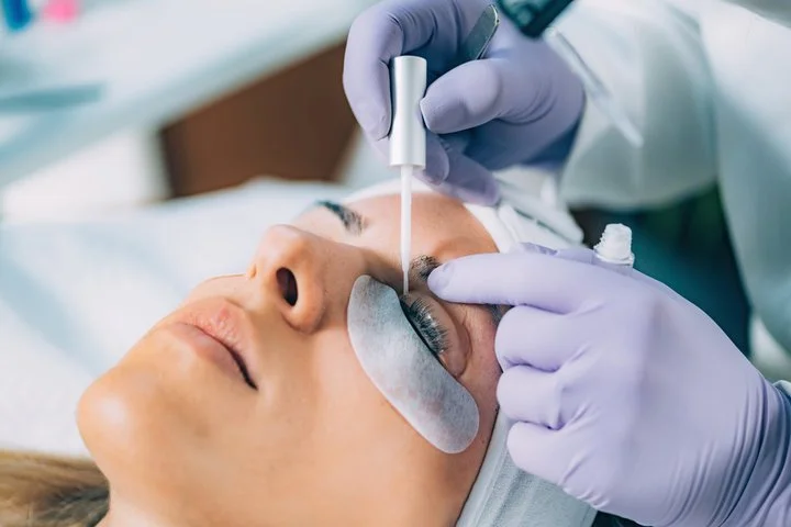 A woman receiving a cosmetic eye treatment with a dropper, covered with eye pads, in a clinical setting.