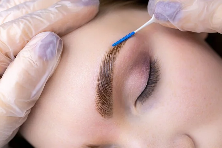 Close-up of a person receiving an eyebrow waxing or grooming treatment with a blue applicator and gloved hands.