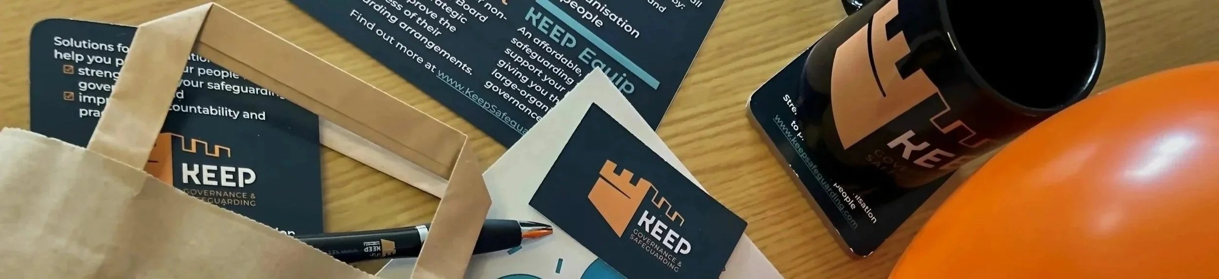 Flyer and merchandise for Keep, a government and community protection organization, featuring a black mug with the Keep logo, two balloons, and promotional materials on a wooden table.