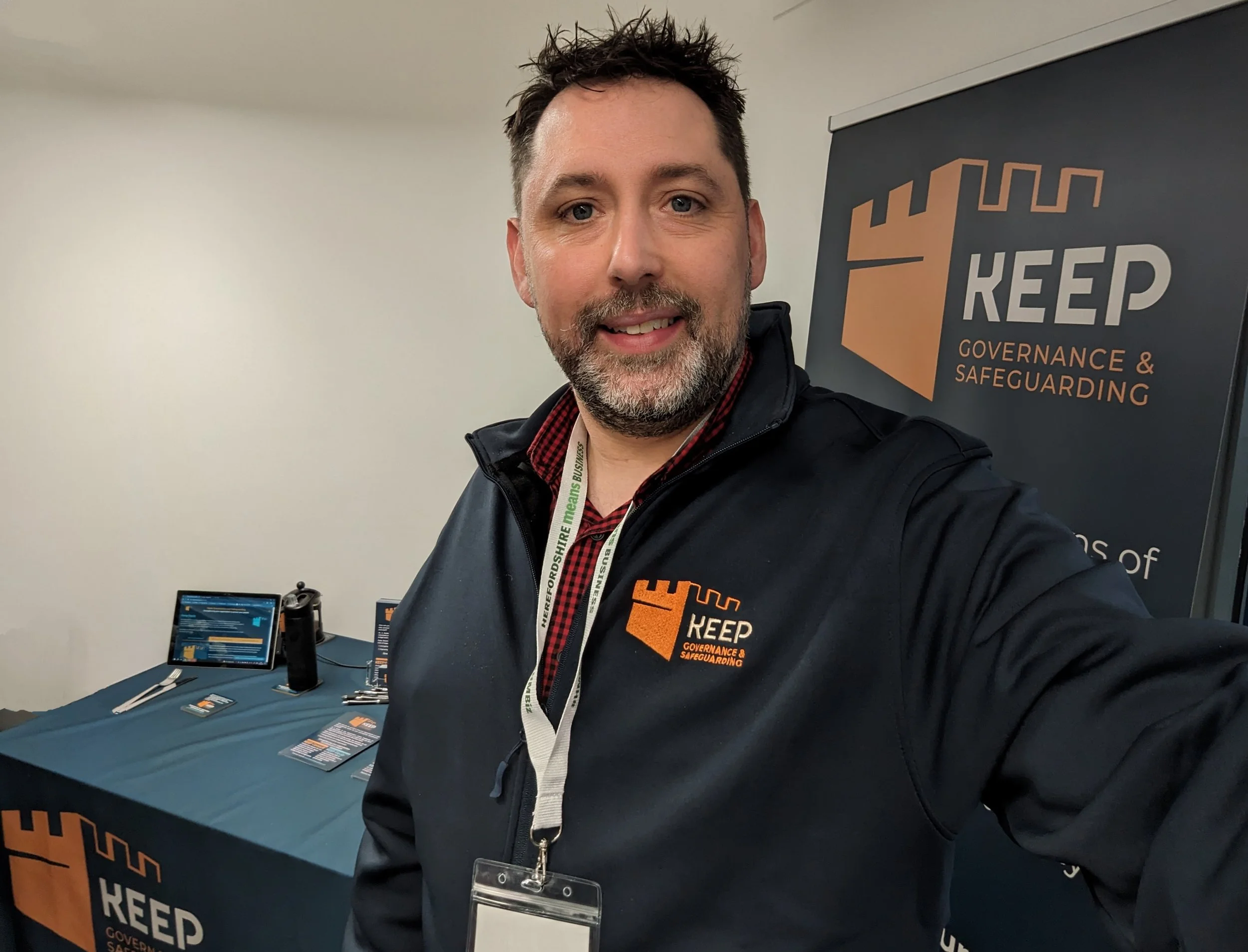 A man with dark, curly hair and a beard taking a selfie at a table with promotional materials for KEEP Governance & Safeguarding. He is wearing a black jacket and a red checkered shirt, with a lanyard around his neck.