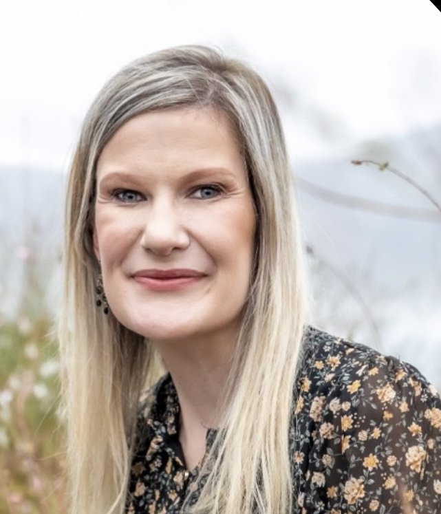 A woman with long blonde hair and blue eyes smiling outdoors, wearing a black floral blouse.