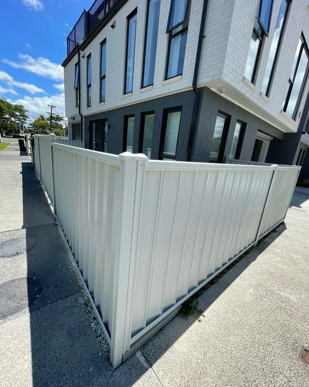 Photo of a modern white metal fence surrounding a small outdoor space in front of a multi-story building with large windows under a blue sky.