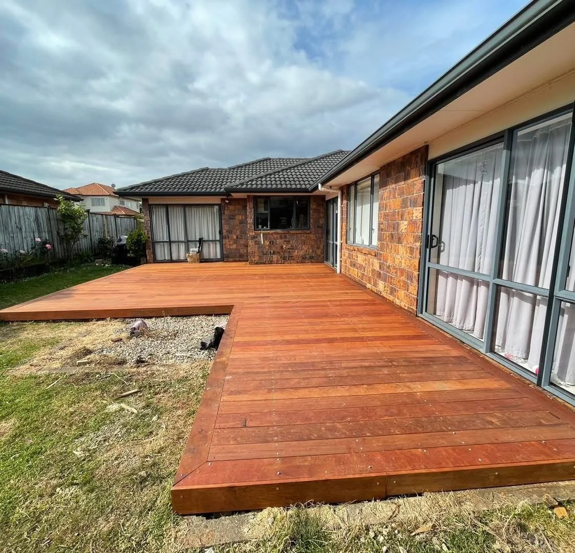 Backyard with newly installed Kwila deck extending from the house, brick exterior walls, windows with white curtains, grassy area to the side, neighboring houses, and cloudy sky overhead.