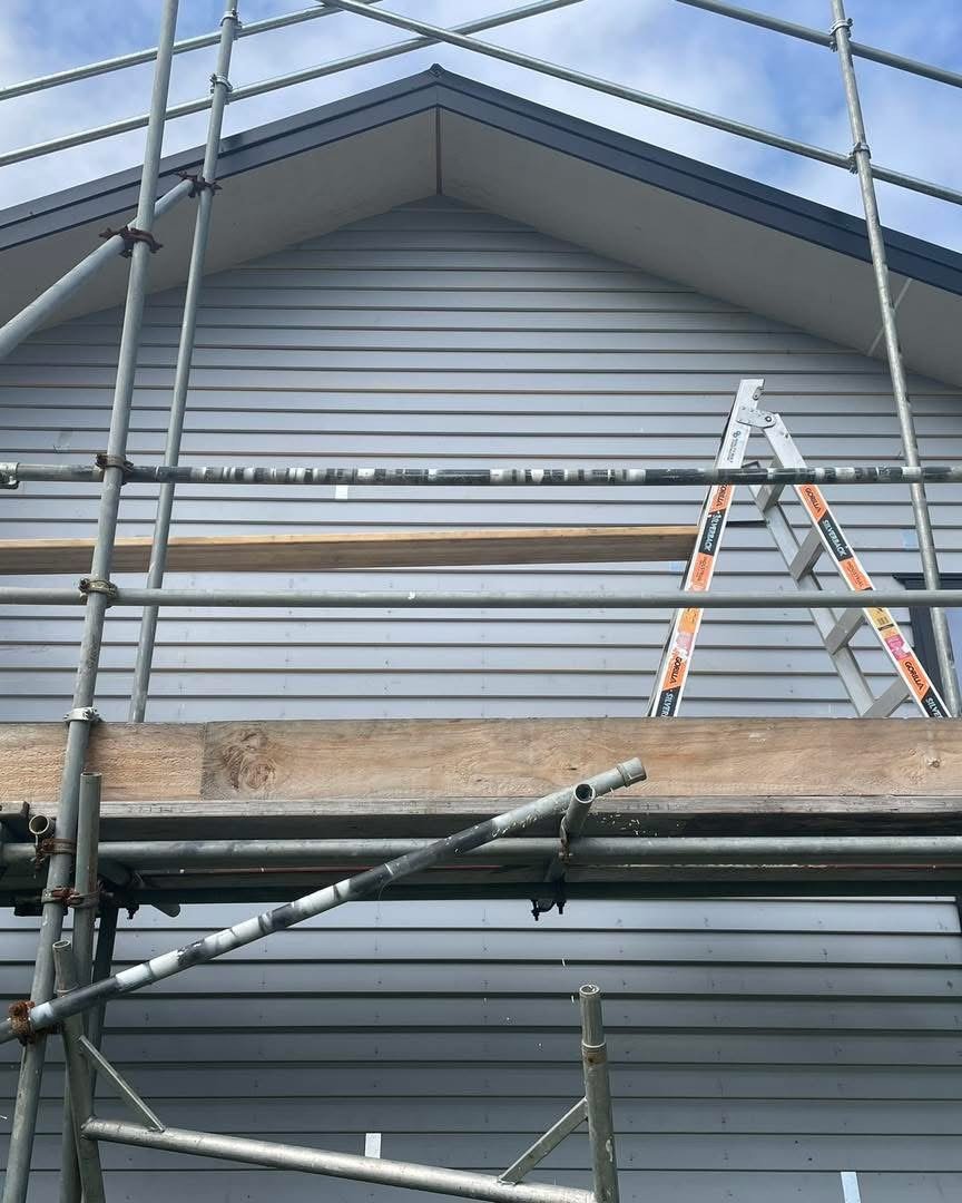 Side view of a house under construction or renovation, with grey siding, a scaffold, and a ladder against the house.