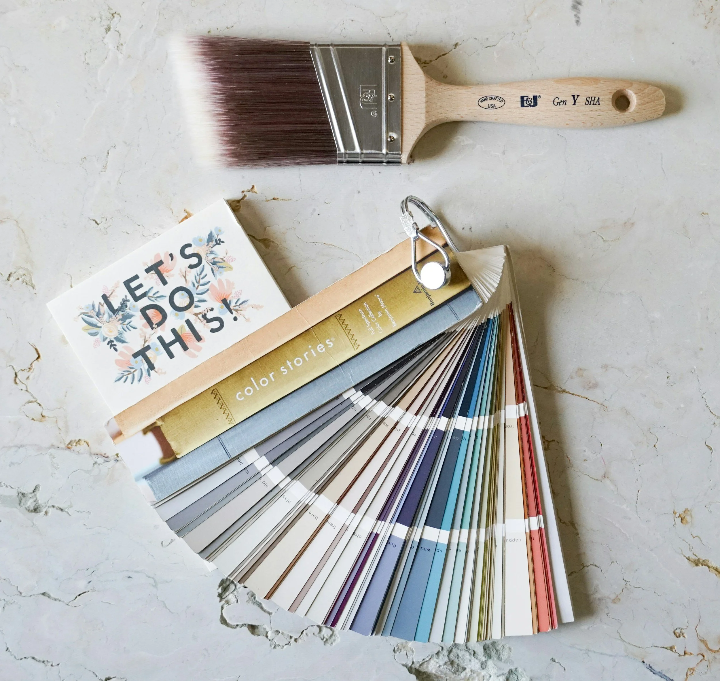 A paintbrush, paint samples, a paint color fan deck, and a small card that says "Let's Do This" with floral illustrations on a marble surface.