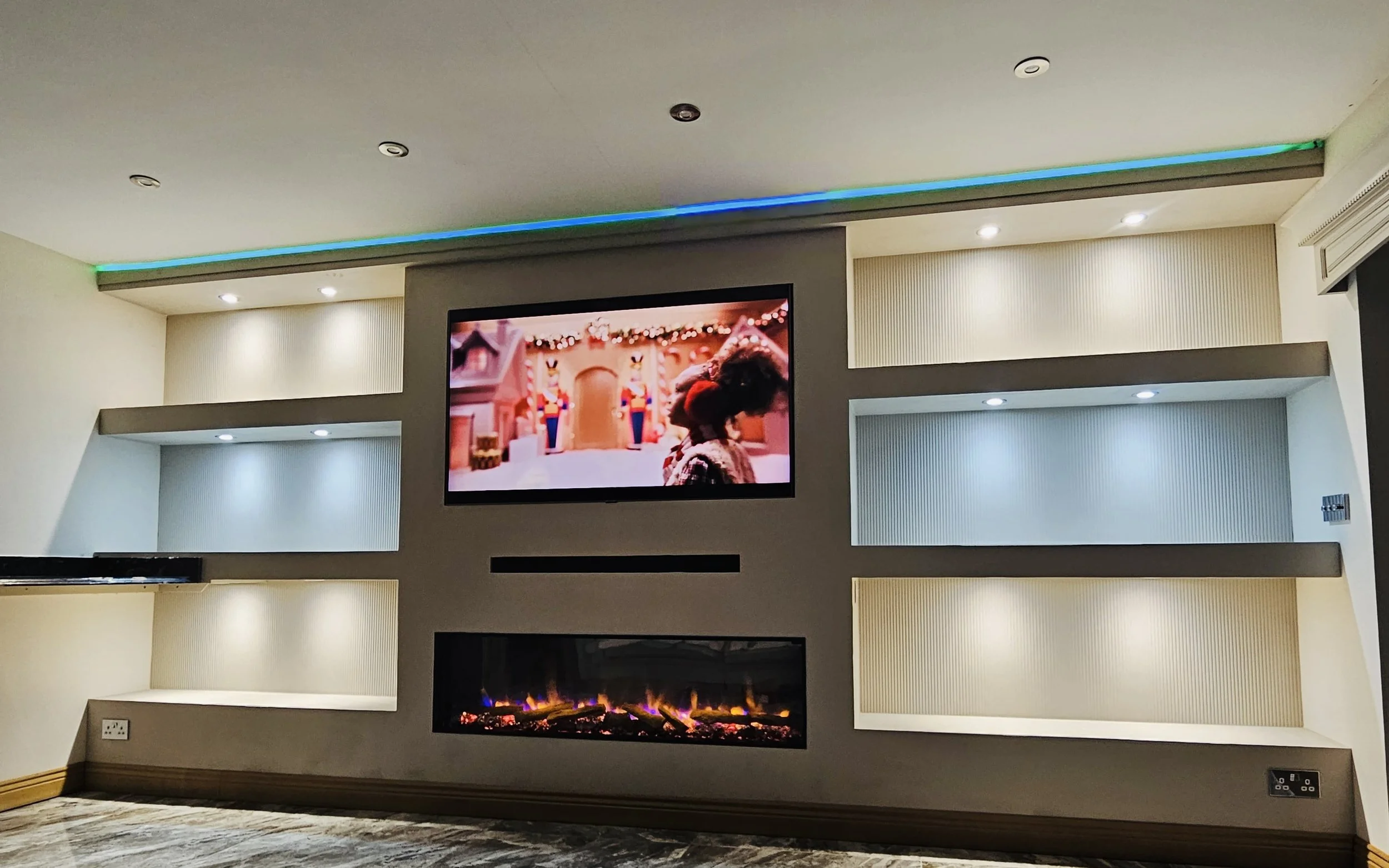 Living room with a mounted flat-screen TV above a modern fireplace with a glass front, surrounded by wooden paneling, next to a side table with a lamp, a cup, and a remote, and a wall with three framed black and white photos.