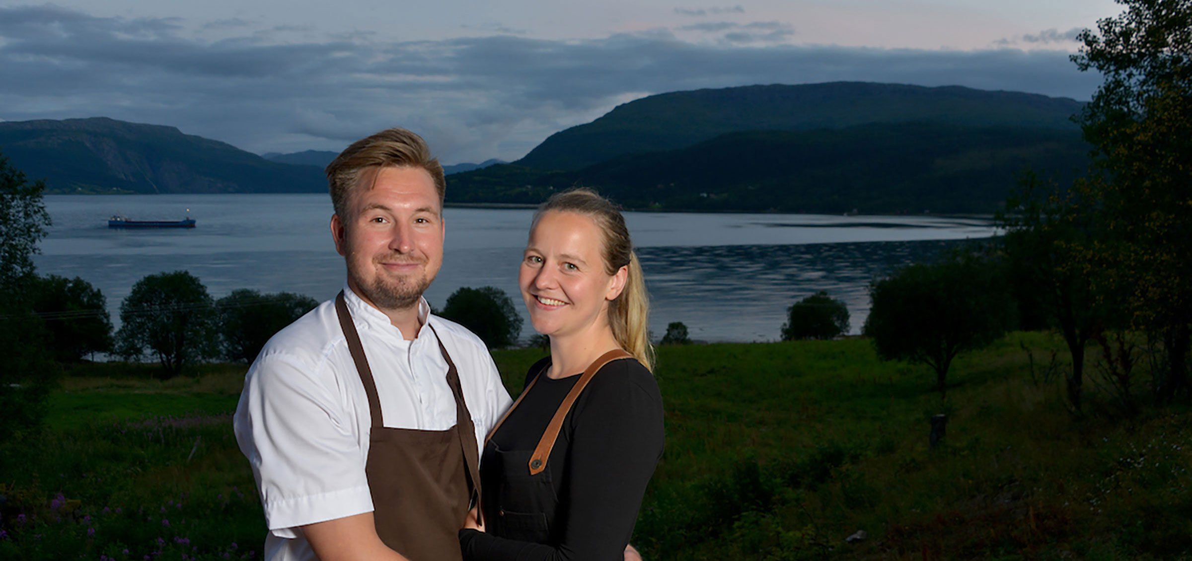 En lykkelig mann og kvinne står sammen med en innsjø og fjell i bakgrunnen ved solnedgang.
