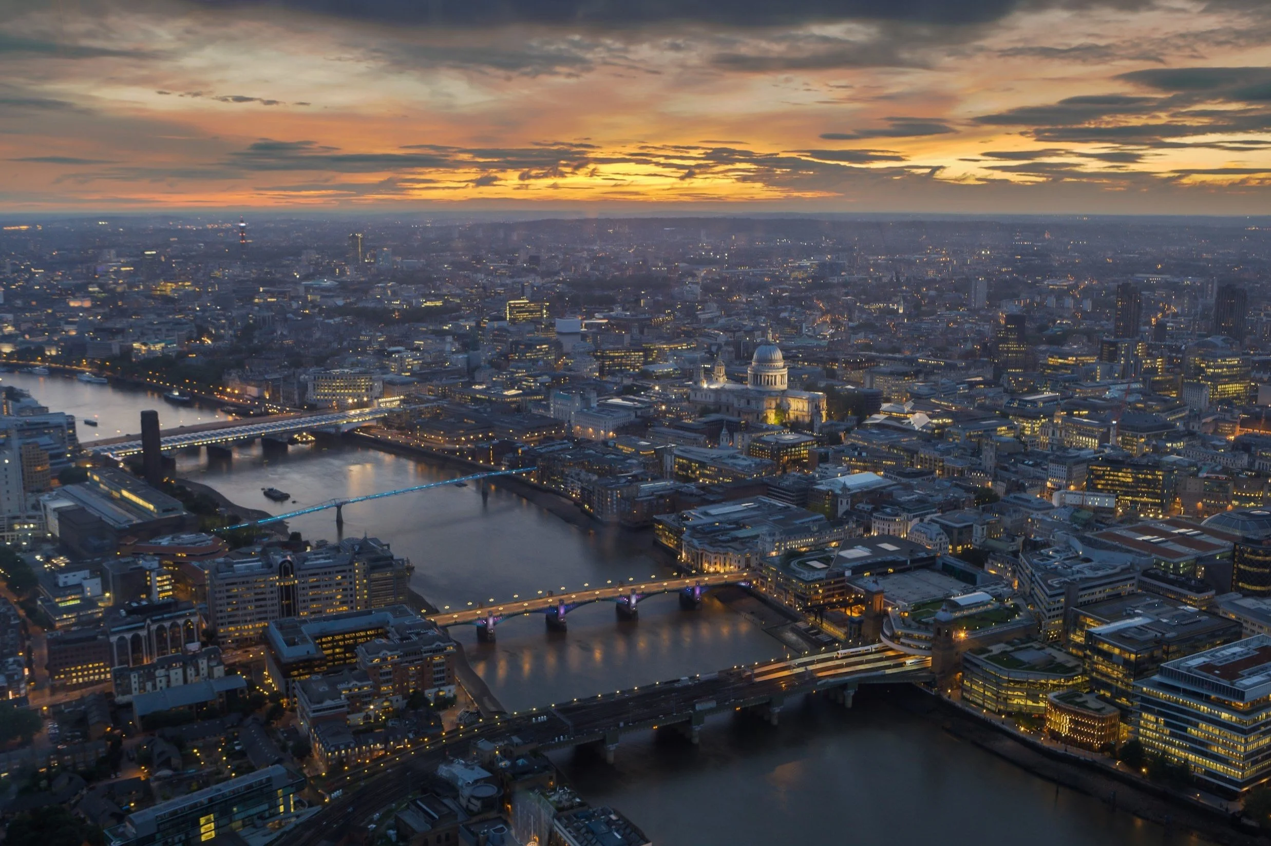 Aerial view of a city at sunset with multiple bridges crossing a river, illuminated buildings, and a domed government building in the skyline.