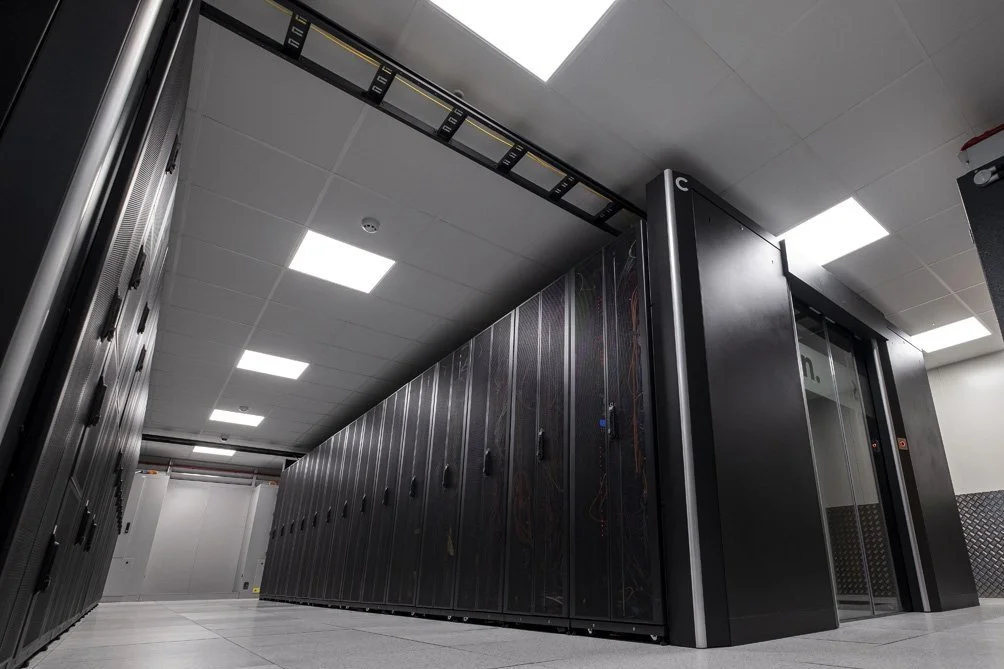 View of a data center server room with black server racks and an elevator door, tiled floor, ceiling lights, and cable management system.