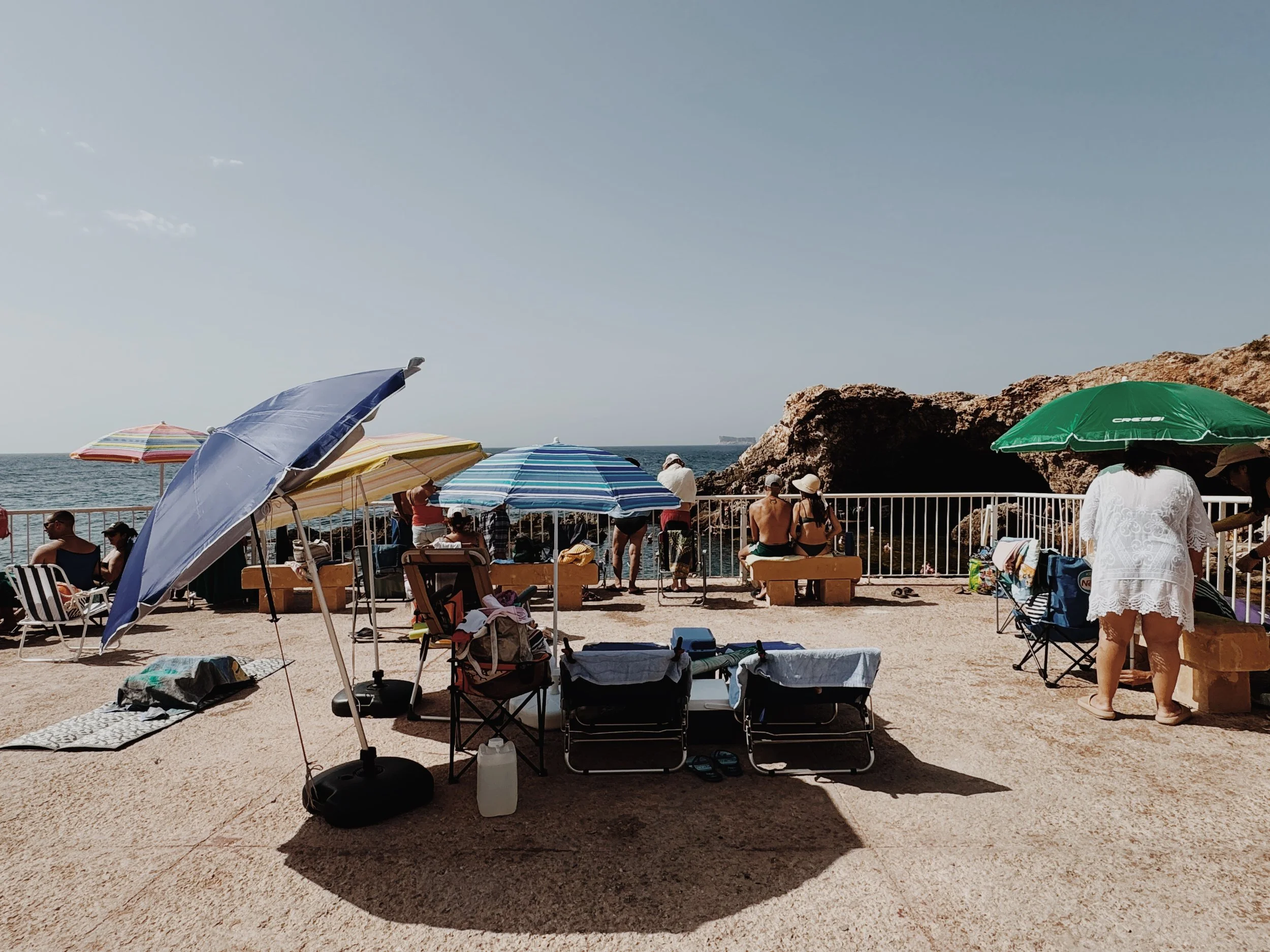 Beach scene with umbrellas, chairs, and people sitting and relaxing by the ocean under clear skies.