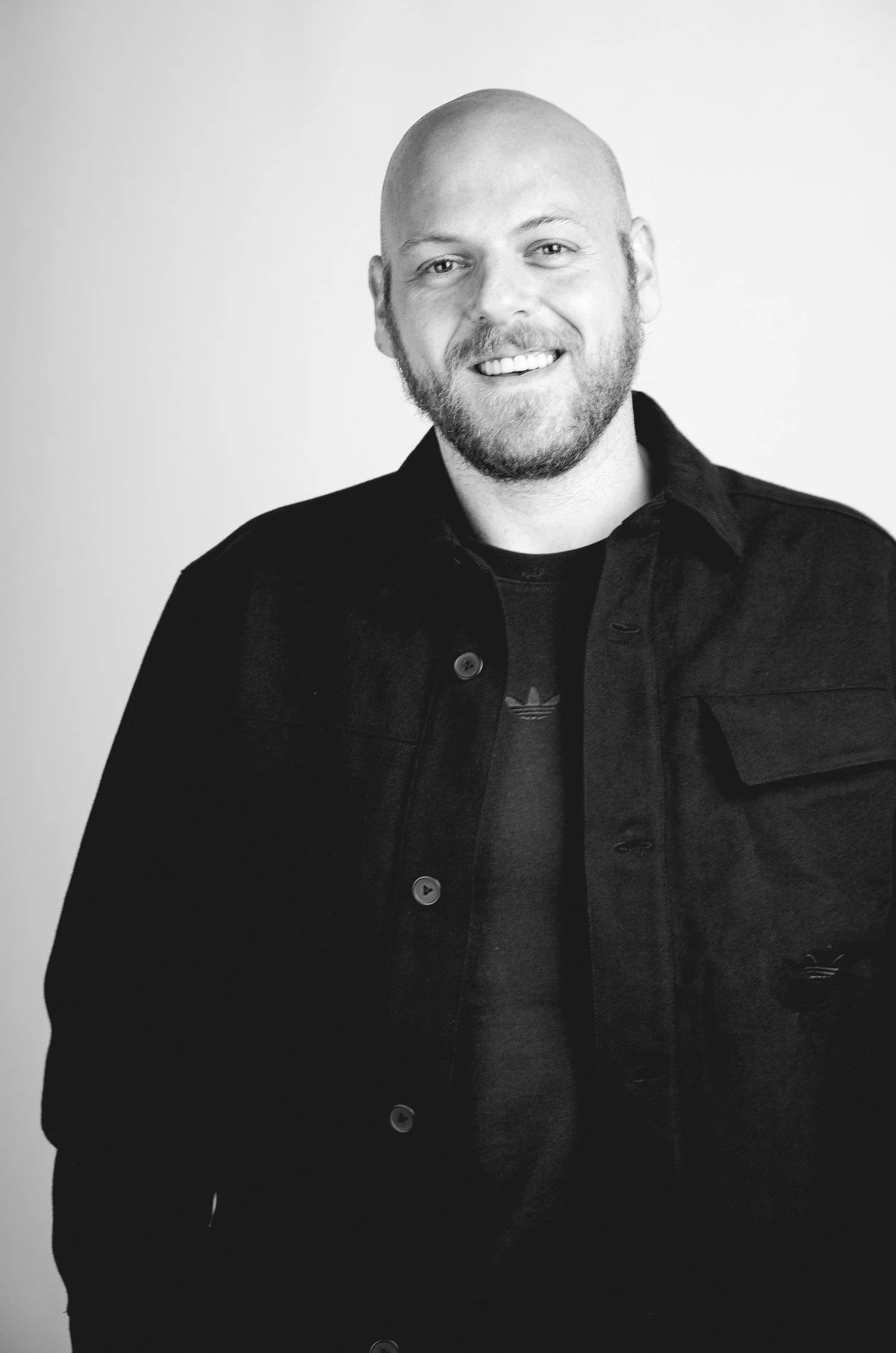 Black and white portrait of a smiling man with a beard, wearing a dark jacket over a T-shirt, standing against a plain background.