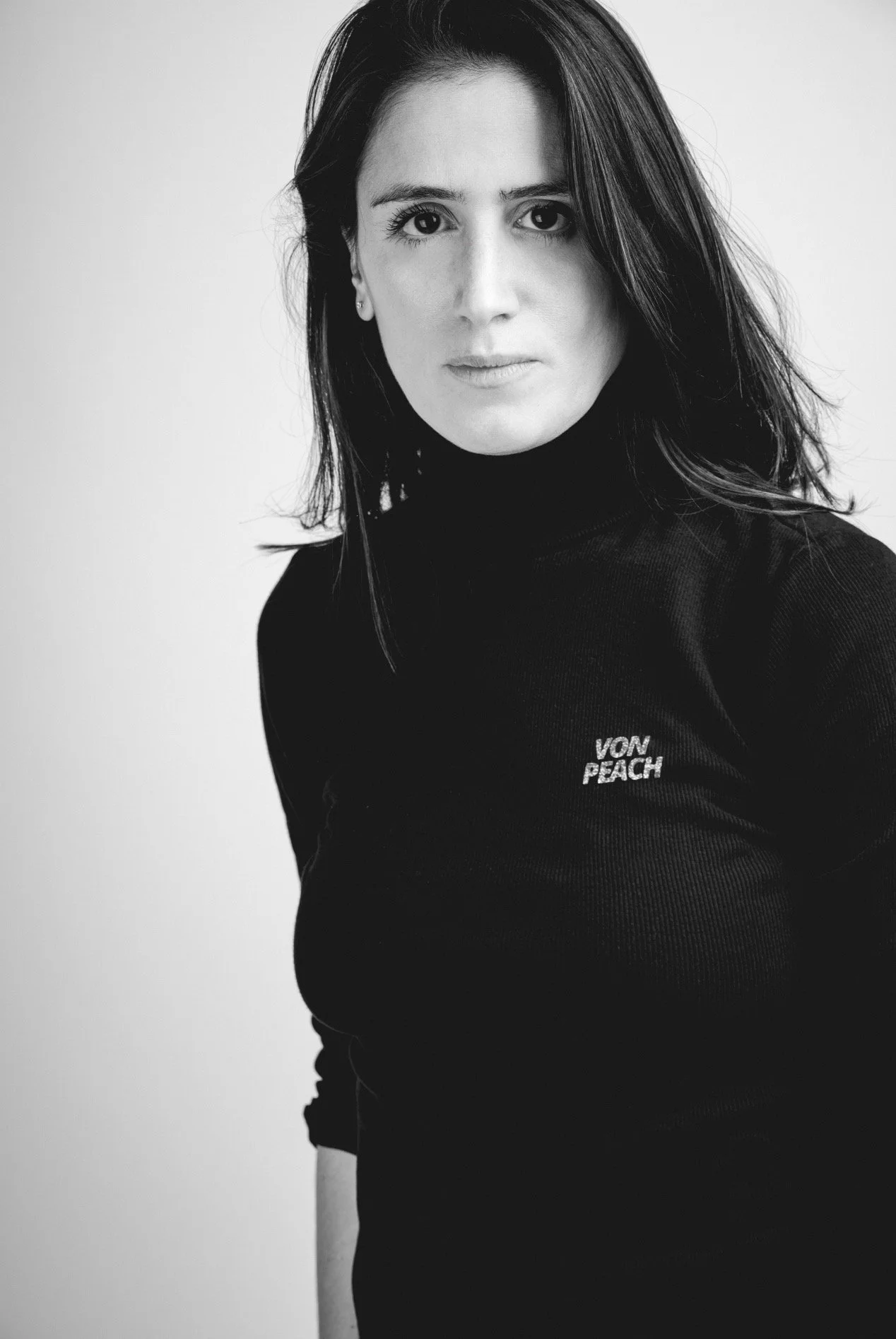Black and white portrait of a woman with shoulder-length dark hair, wearing a black turtleneck with "VON PEACH" embroidered on it, looking directly at the camera.