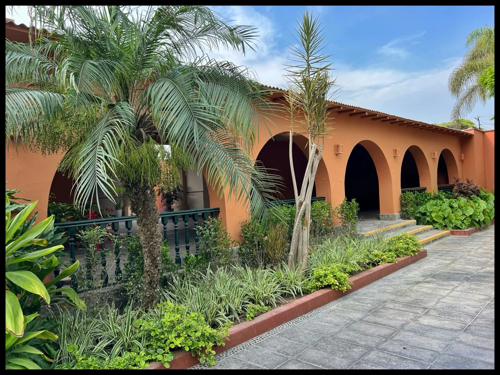 Edificio de color terracota con arcos en una entrada, rodeado de plantas verdes y palmeras, en un día soleado con cielo azul y algunas nubes.