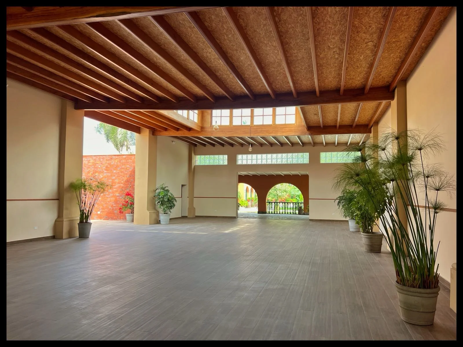 Espacio interior amplio con paredes claras, suelo de baldosas y techo de madera con vigas expuestas, decorado con plantas en macetas grandes.