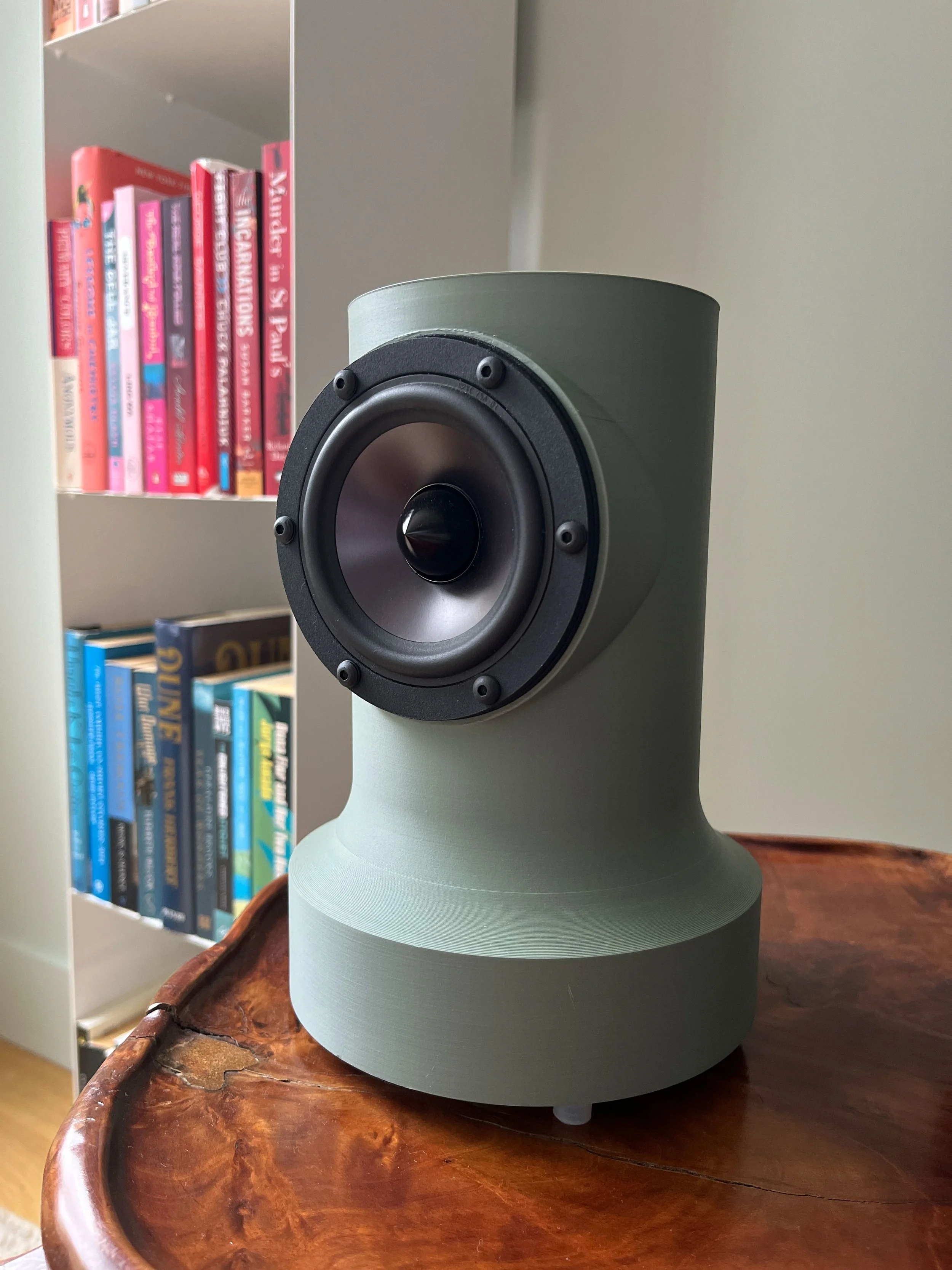 A modern speaker with a gray casing and black speaker cone, placed on a wooden surface with bookshelves in the background.
