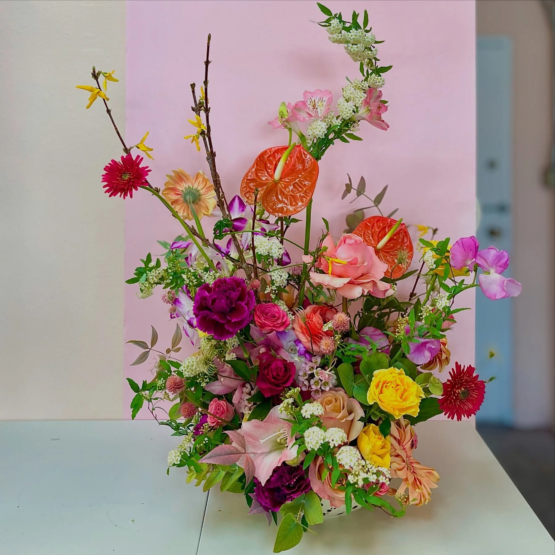 Colorful bouquet of various flowers including pink roses, purple carnations, yellow daisies, white baby's breath, and anthuriums, with branches and green leaves, arranged on a white surface with a pink and white background.