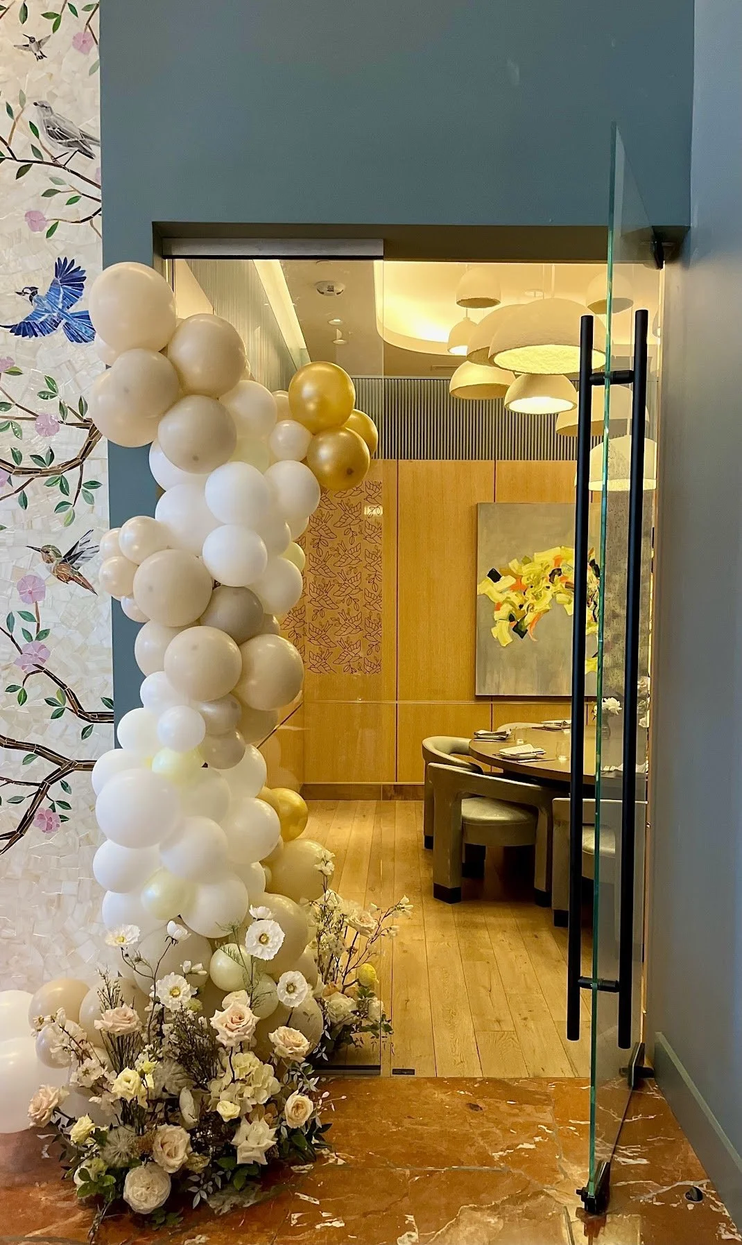 Decorative entrance with balloon arch and floral arrangement leading into a room with wooden decor, modern lighting, and colorful artwork on the wall.