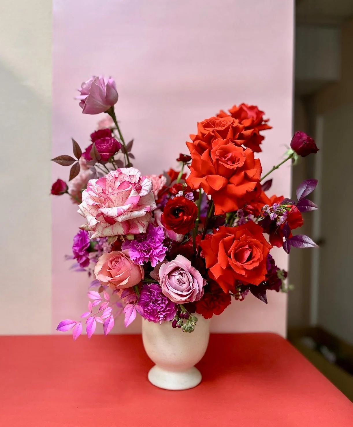 Colorful floral arrangement with roses, carnations, and other flowers in a white vase on a red table, with a pink background.