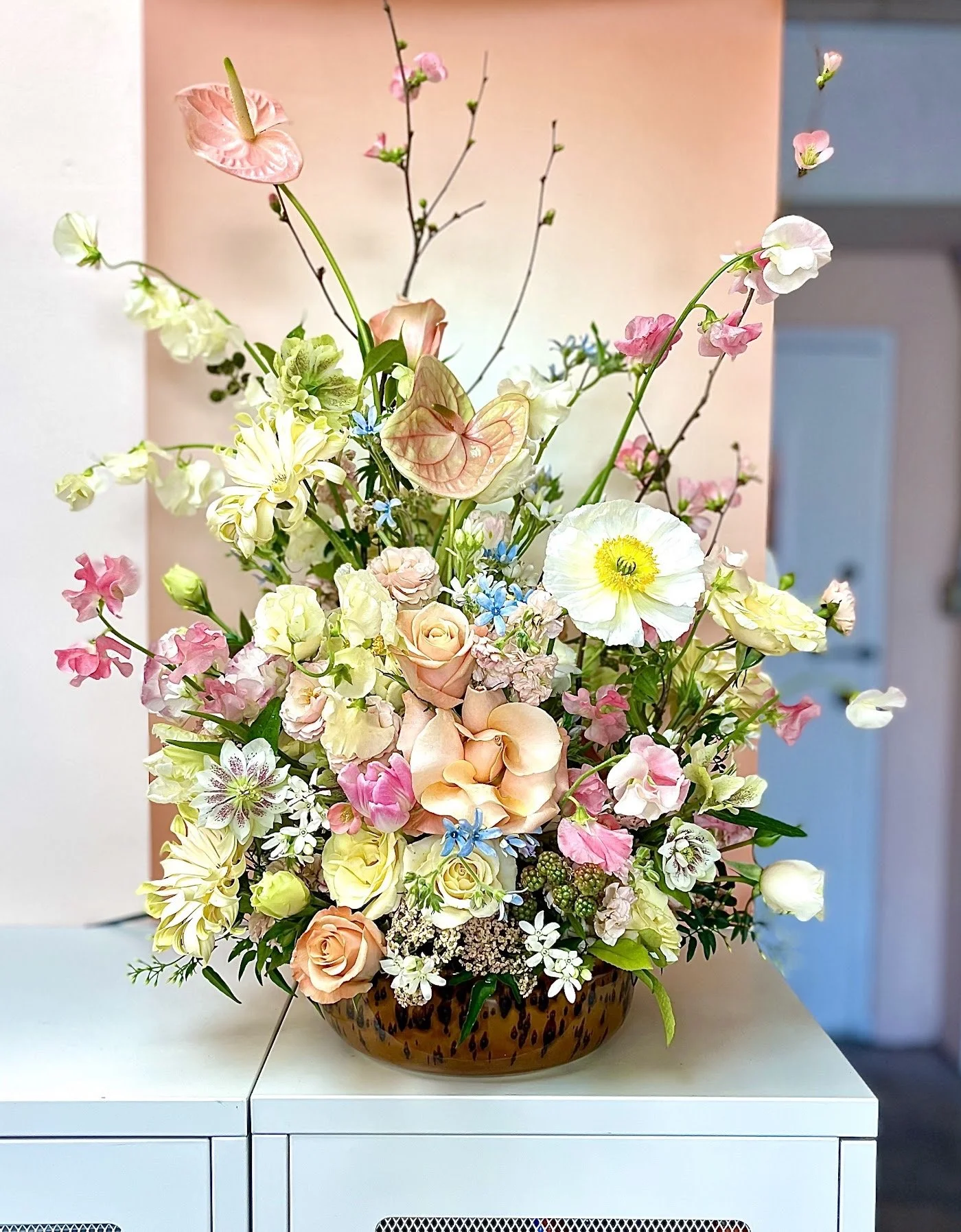 A colorful flower arrangement in a brown, oval-shaped vase on a white surface, featuring roses, daisies, jasmine, and other mixed flowers in pastel shades of pink, white, peach, and blue, with some branches and greenery.