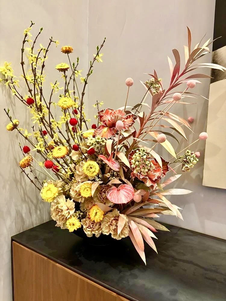 A colorful flower arrangement with yellow, red, pink, and cream flowers, including branches with small pink pom-pom like flowers, displayed on a black surface against a neutral wall.
