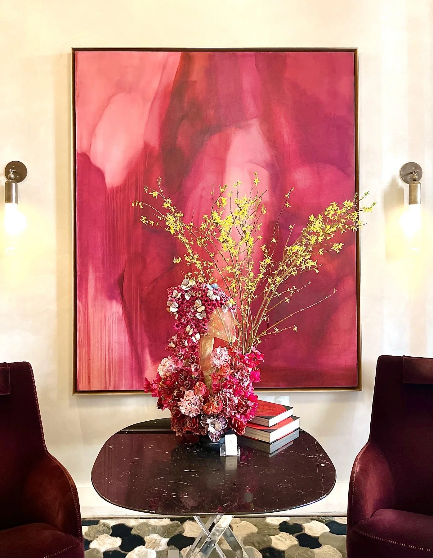 Decorative flower arrangement with pink and white flowers and green branches on a black table, with an abstract pink and red painting on the wall behind, flanked by two wall-mounted lights, and two burgundy armchairs on either side.