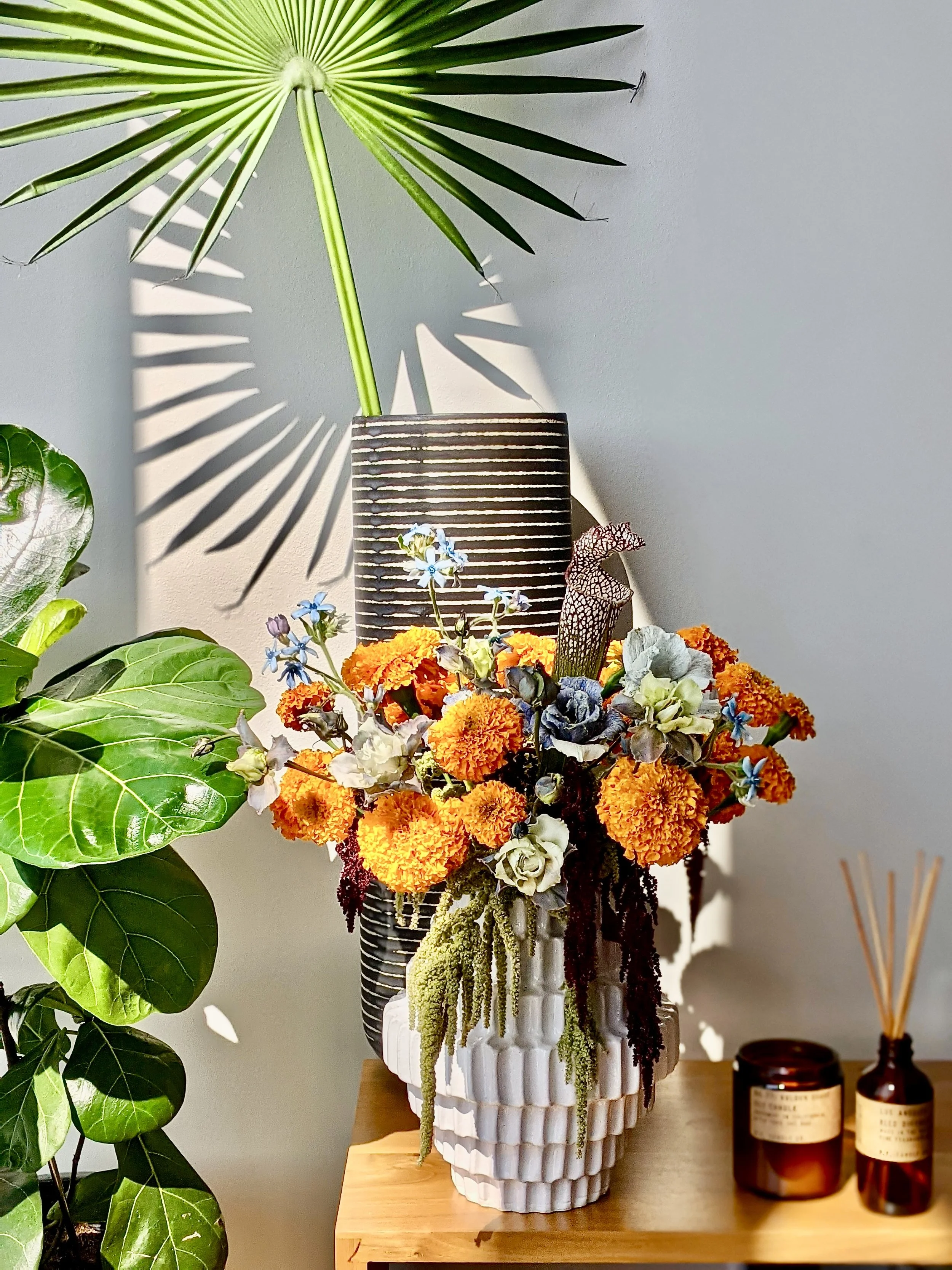 A white textured vase with a variety of colorful flowers including orange marigolds, purple, white, and blue blooms. Behind it, a black and white striped vase holds a large green palm leaf with distinct shadow patterns on the wall.