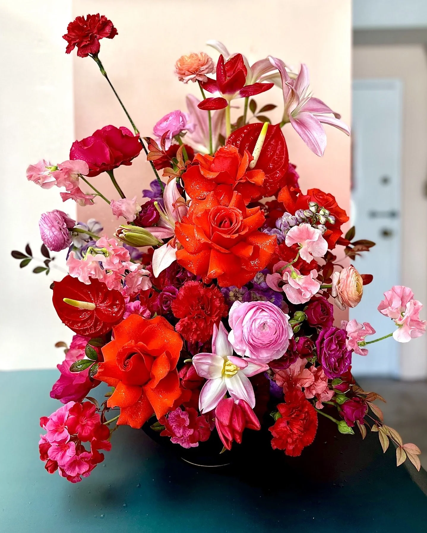 A vibrant bouquet of various red, pink, and purple flowers arranged in a black vase, placed on a teal surface with a beige wall and a door in the background.