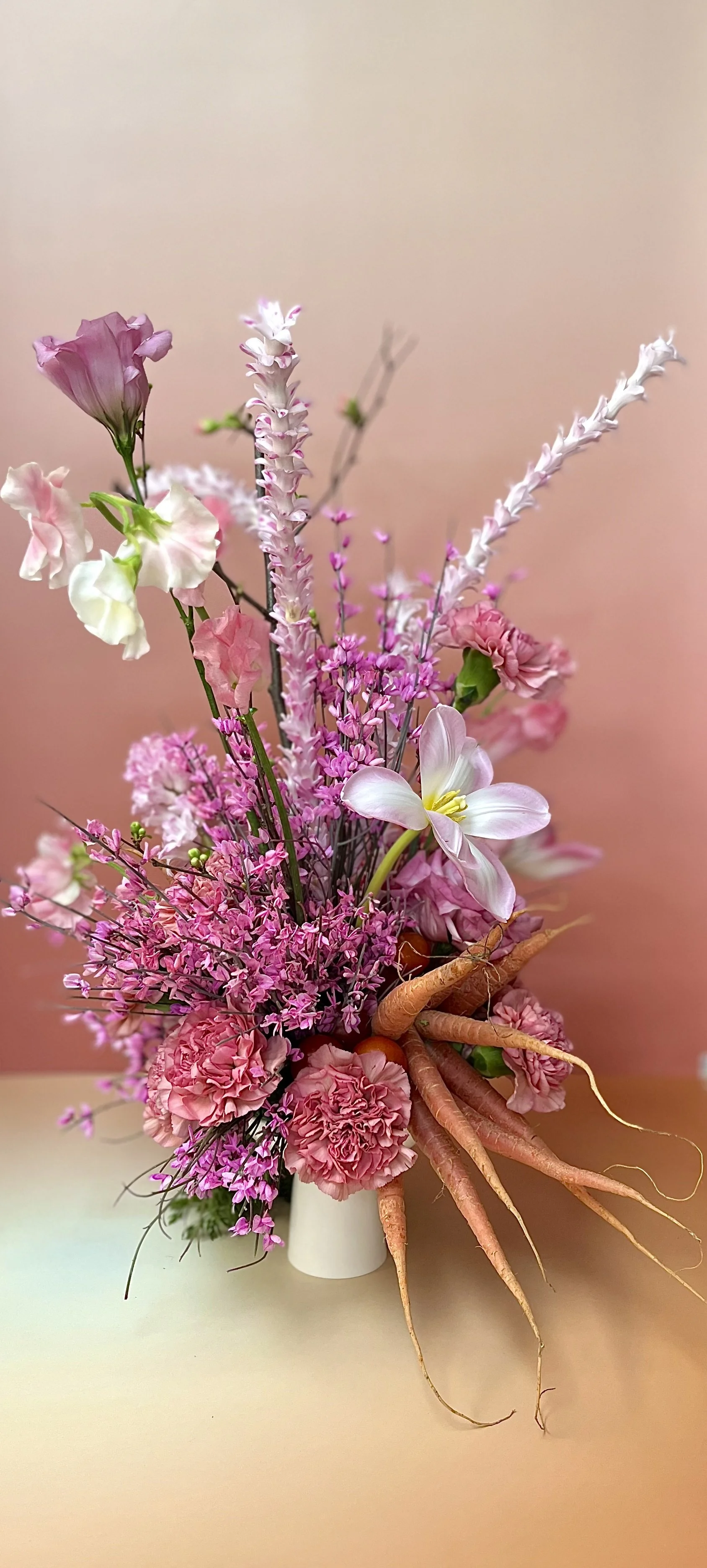 Arrangement of pink, white, and purple flowers with roots displayed in a white vase against a pink background.