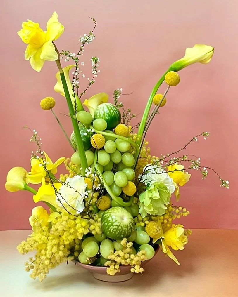 Arrangement of yellow flowers, green grapes, miniature watermelons, small white flowers, and branches in a pink vase against a pink background.
