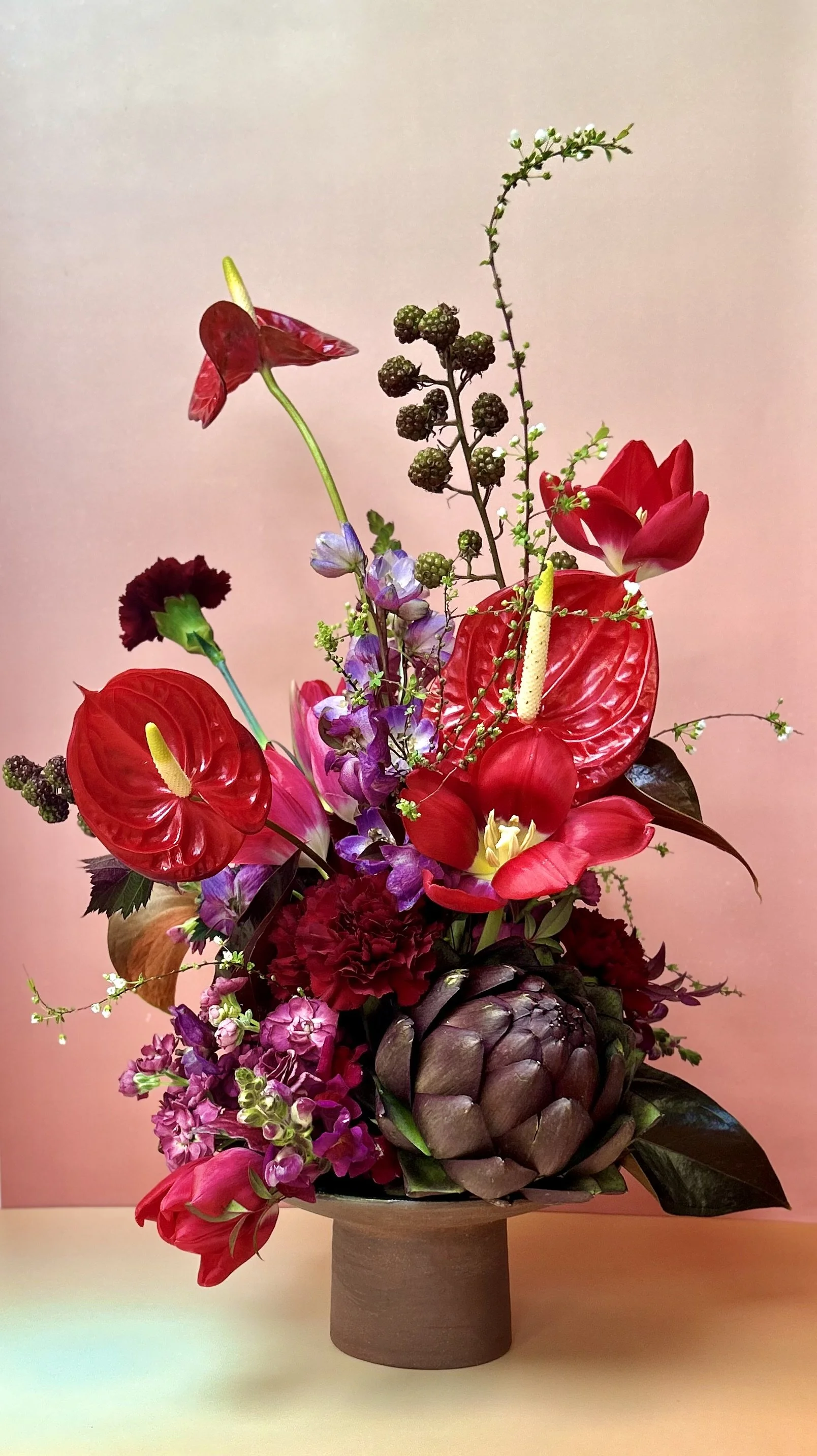 A colorful flower arrangement in a dark brown vase, featuring red anthuriums, pink tulips, purple hyacinths, and green foliage, set against a pink background.