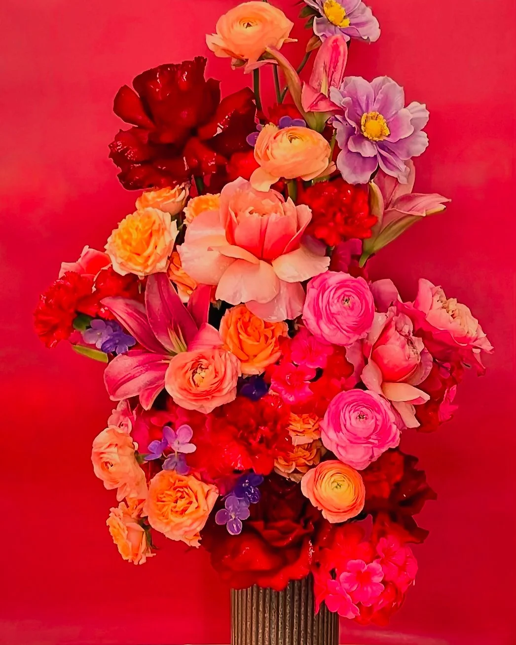 Colorful bouquet of various flowers including roses, lilies, carnations, and other blossoms in pink, red, purple, and peach tones set against a pink background.