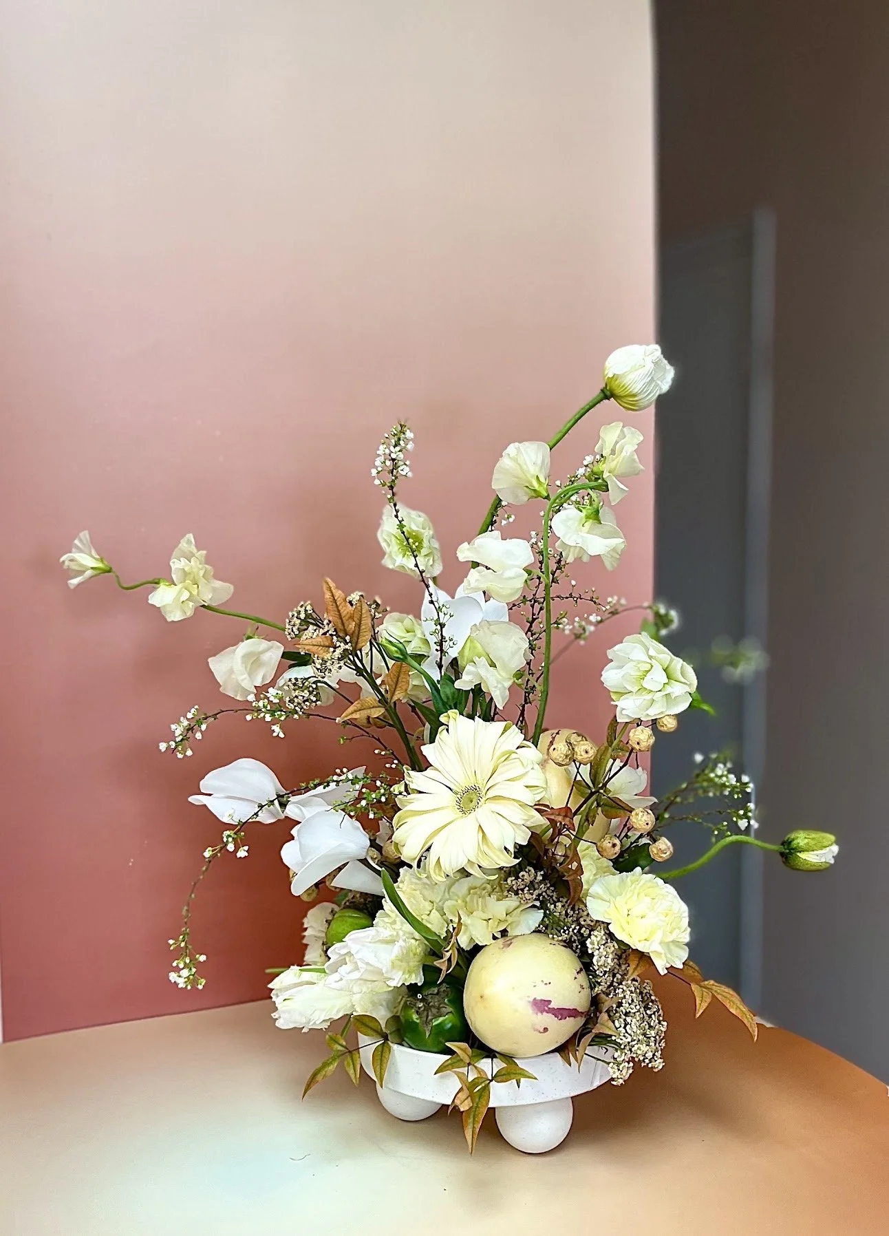 A floral arrangement with white and cream-colored flowers, leaves, and an apple in a white vase on a table with a pink and gray background.