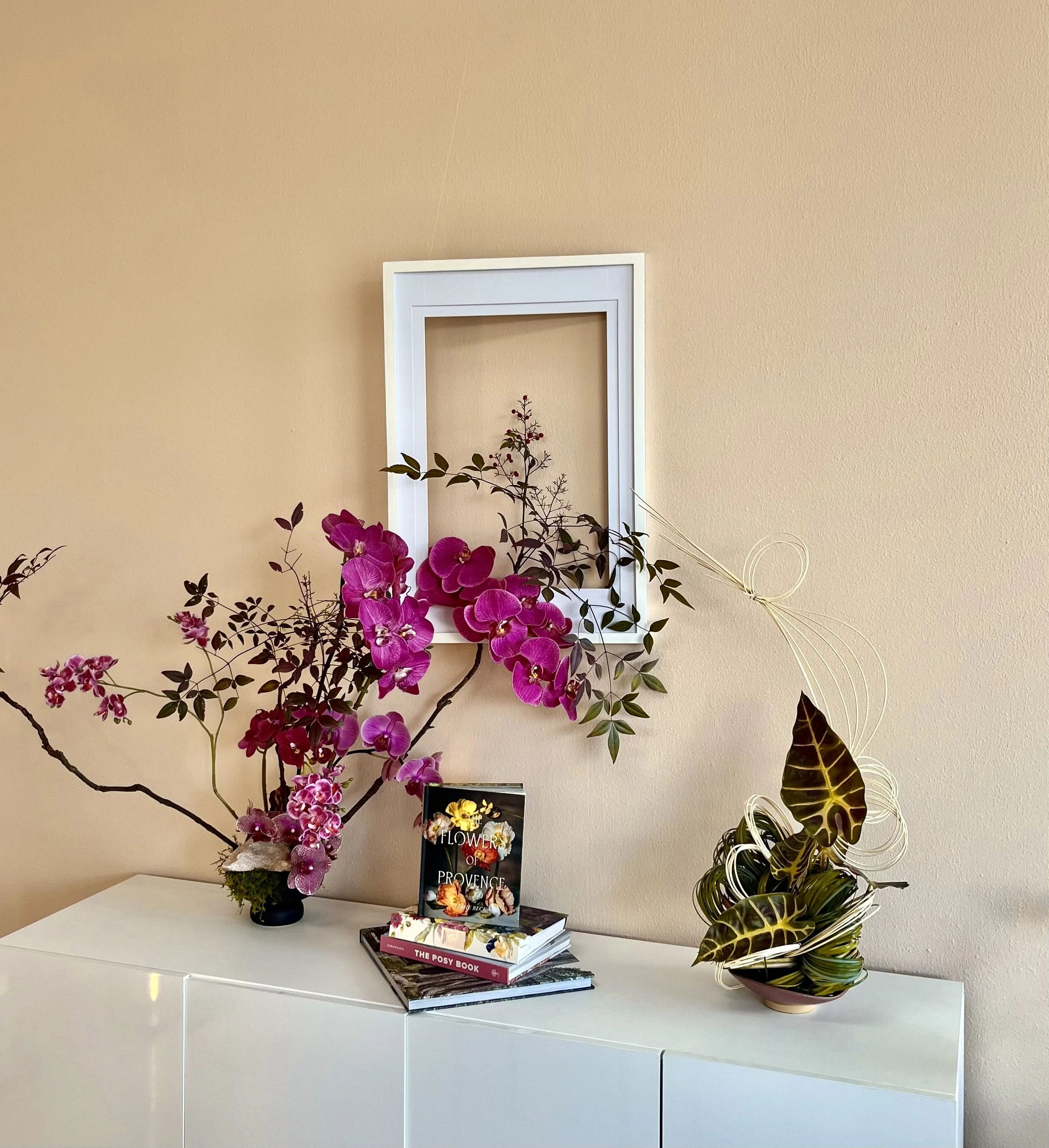 Pink orchid flower arrangement on a white cabinet with books and a plastic pot with large dark green and yellow leaves, beige wall, and a white empty picture frame in the background.