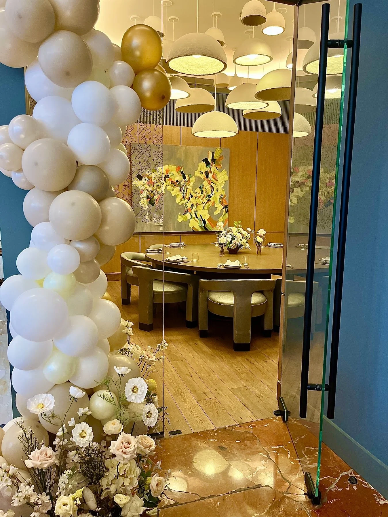 A decorated dining room seen through an open glass door, featuring a round table with floral arrangements, surrounded by chairs, with balloons and artwork on the walls.