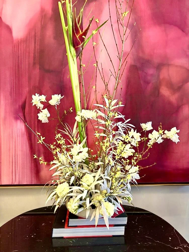 White floral arrangement with tall green stalks and branches, placed on top of stacked books in front of a red abstract background.