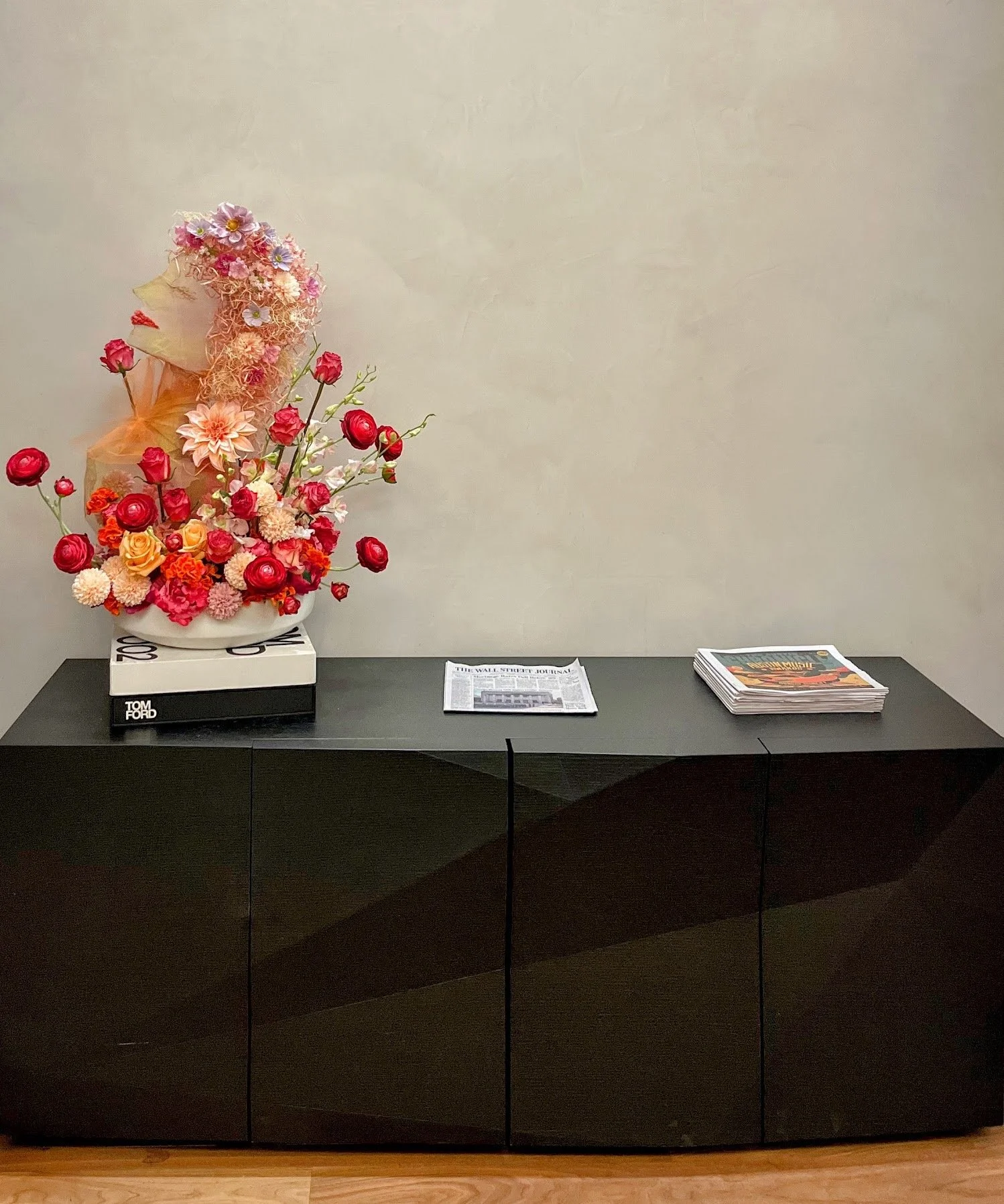 Decorative floral sculpture with pink roses, peach and pink flowers, arranged on a black cabinet, with newspapers and magazines nearby.