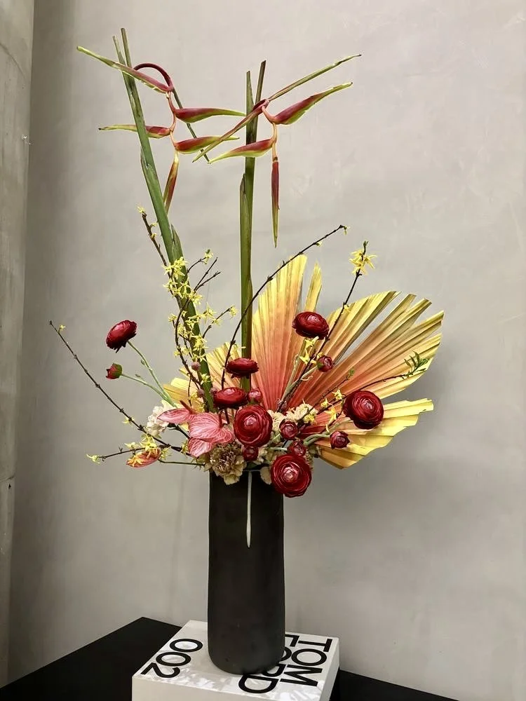 A tall black vase with an artistic arrangement of flowers and leaves, including red roses, pink flowers, yellow blossoms, green leaves, and a dried yellow palm leaf, placed on a white pedestal against a plain wall.