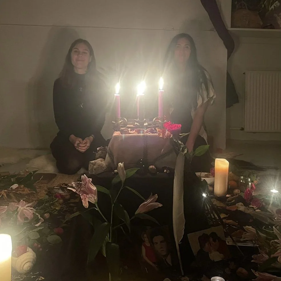 Two women sit on the floor behind a altar with lit candles, flowers, and photographs, creating a memorial setup.