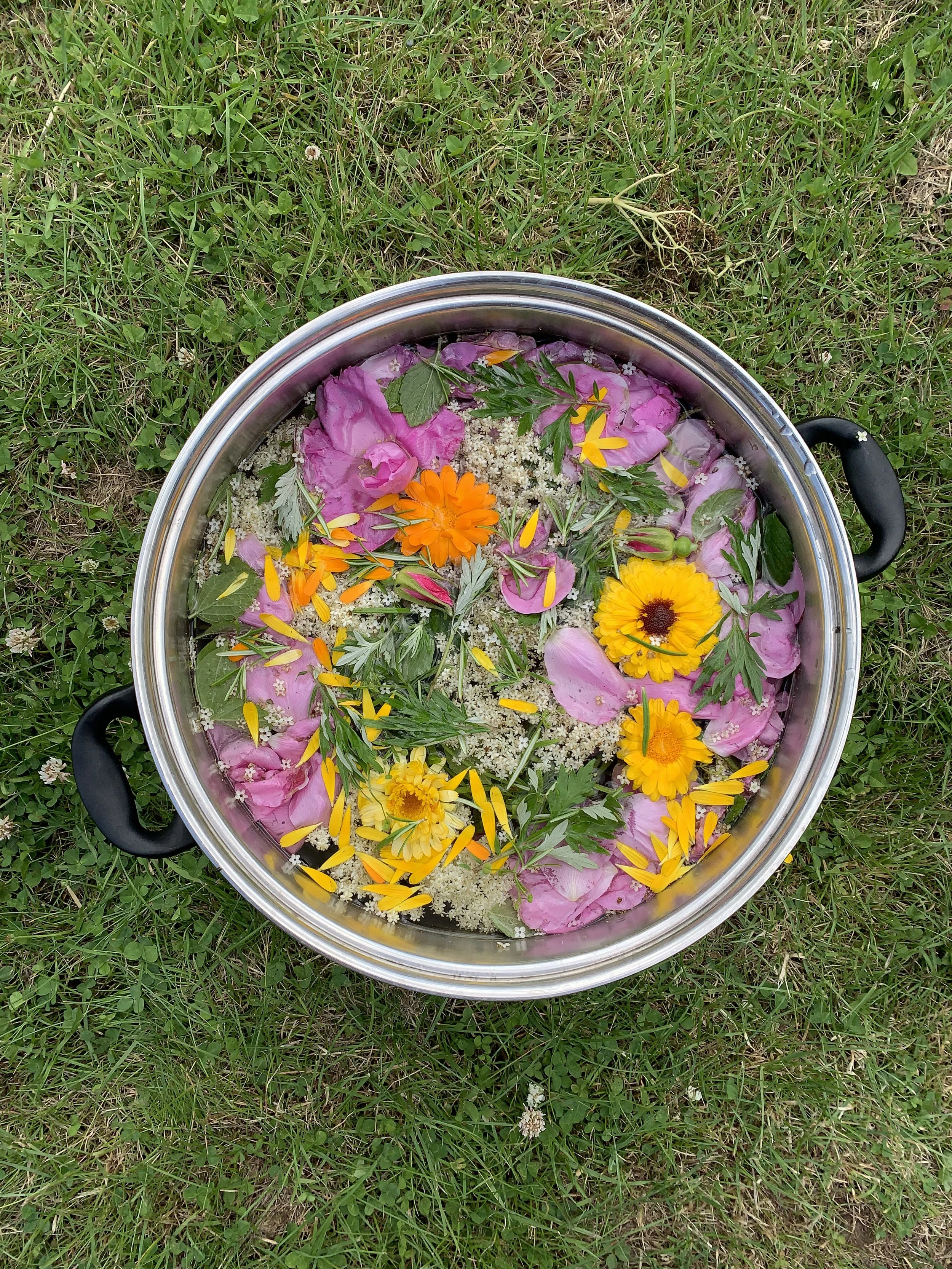 A metal basin filled with water, decorated with colorful flower petals and real flowers such as marigolds and daisies, resting on grass. For a flower bath. 