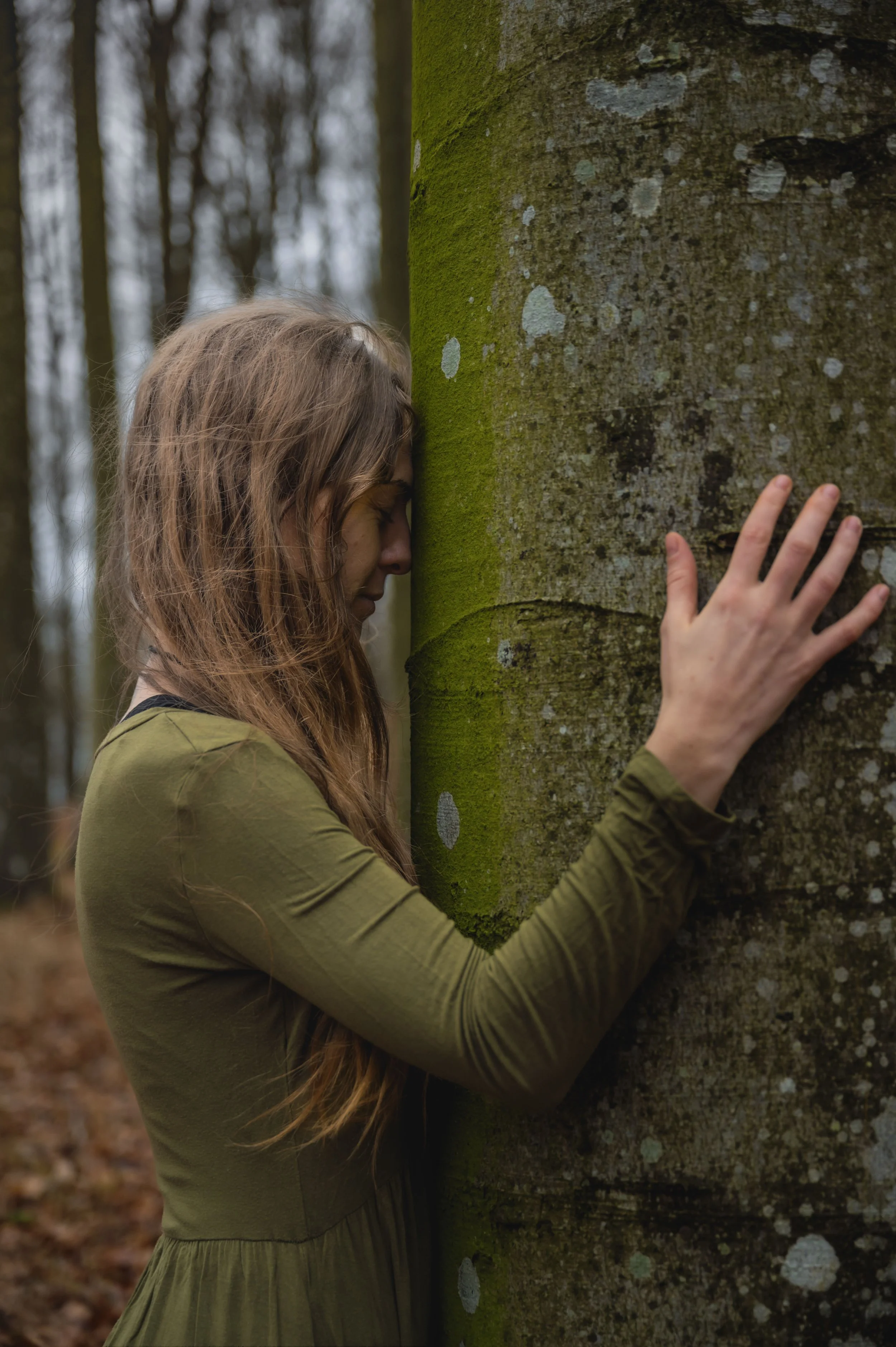A young woman with long hair rests her forehead and hand on a moss-covered tree trunk in a forest, with her eyes closed and a peaceful expression.