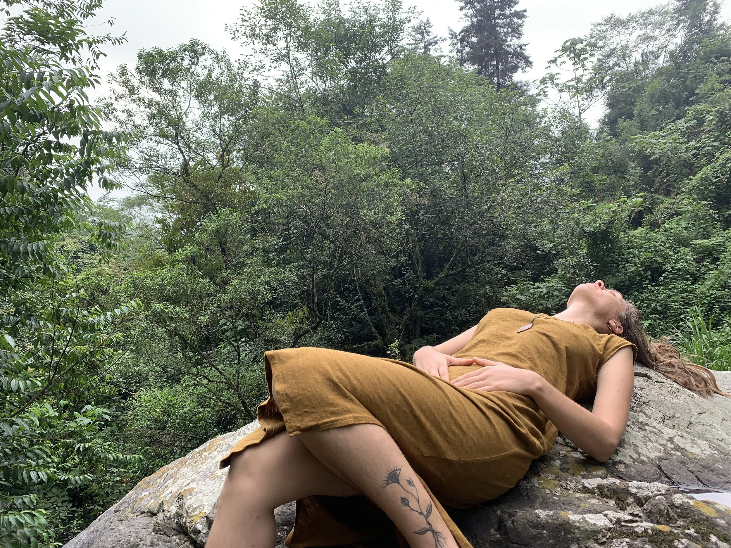 Kaya lying on a large rock in a lush green forest, touching her womb space.