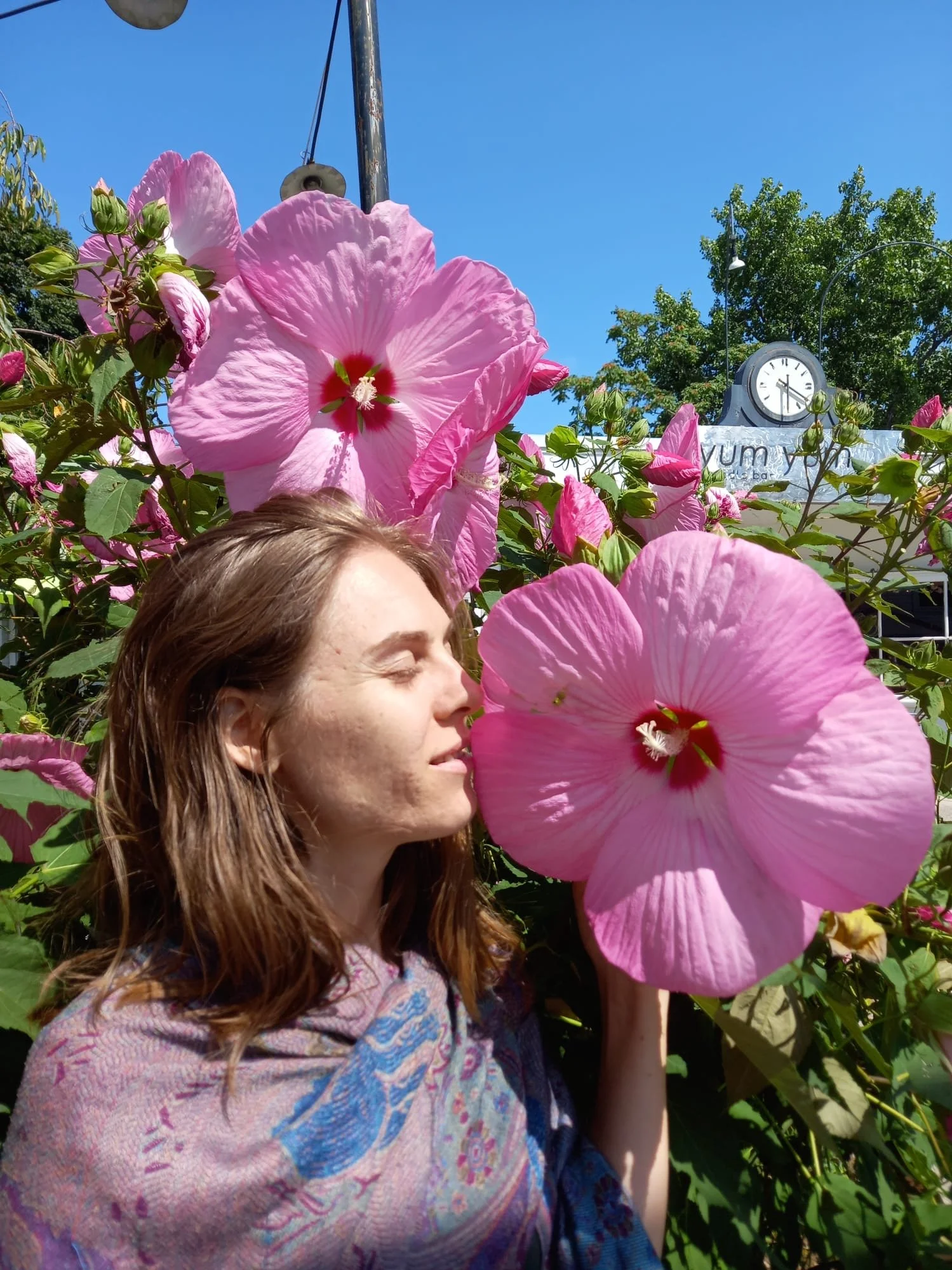 Kaya with closed eyes and smiling, smelling pink hibiscus flowers in a garden on a sunny day with a blue sky and clock in the background.
