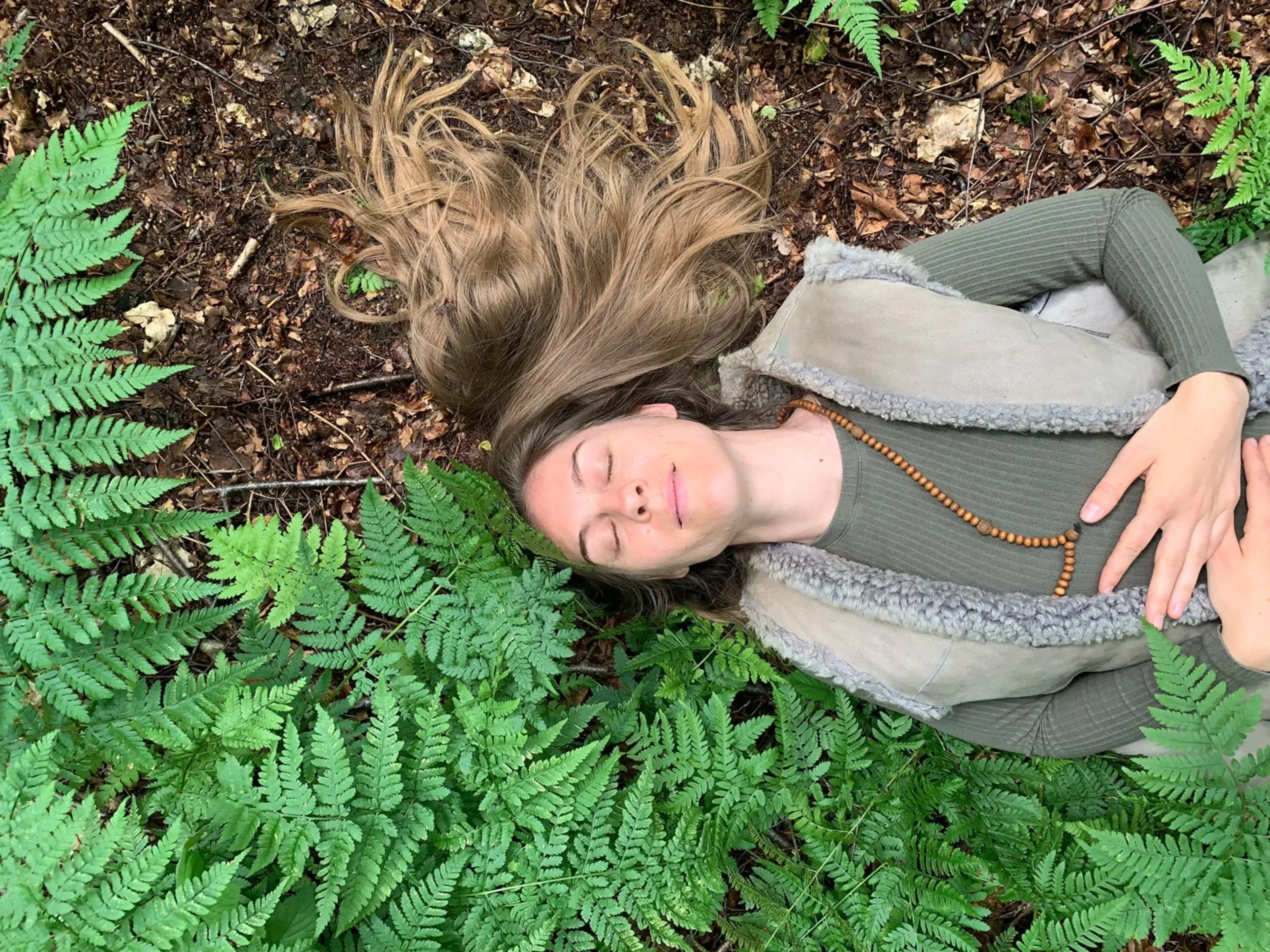 Kaya lies on the forest ground amidst green ferns and brown leaves, eyes closed and hands on her stomach, appearing peaceful and relaxed.