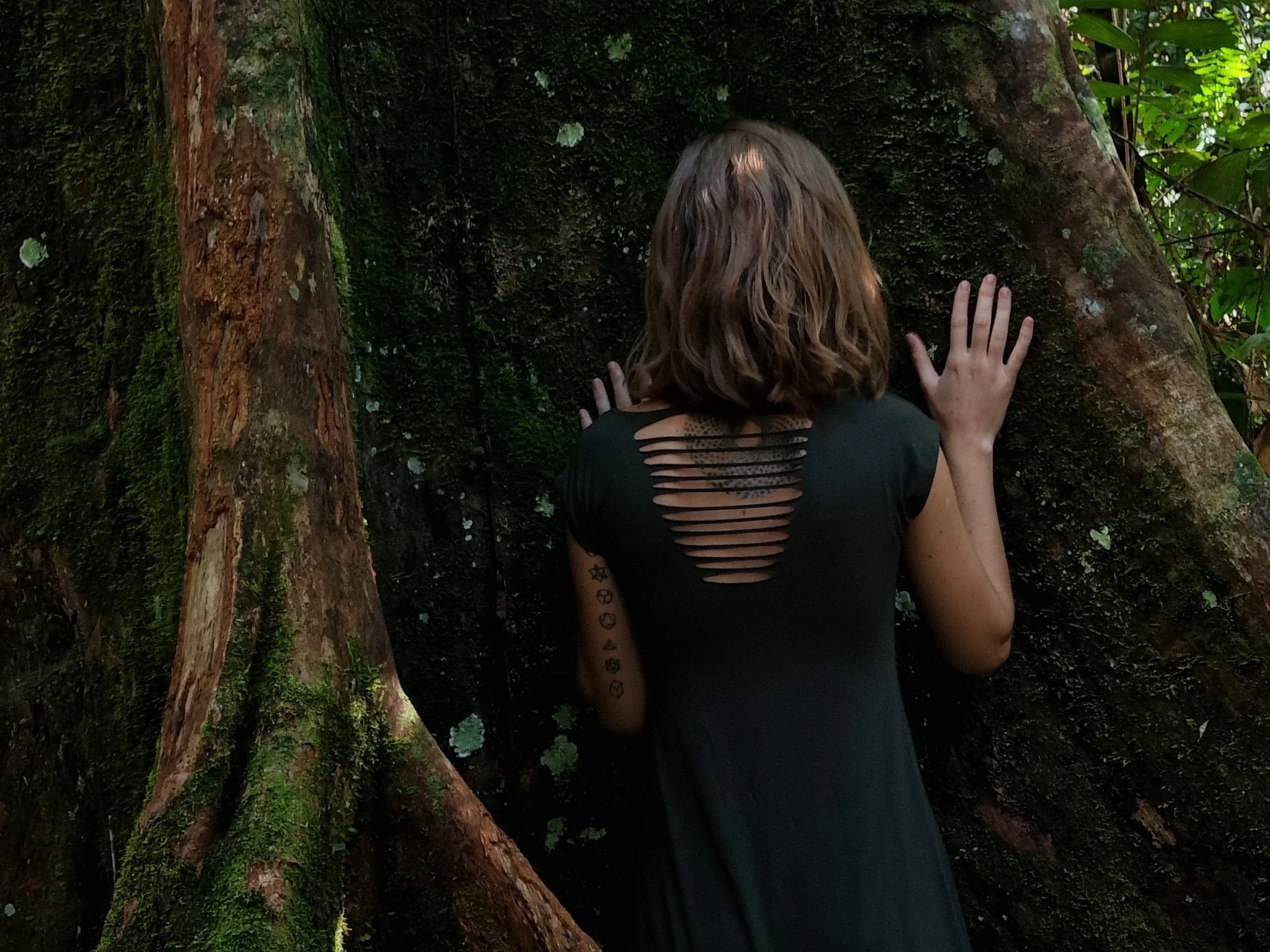 Kaya stands facing a large tree with moss and lichen, touching the tree with both hands in a lush green forest.