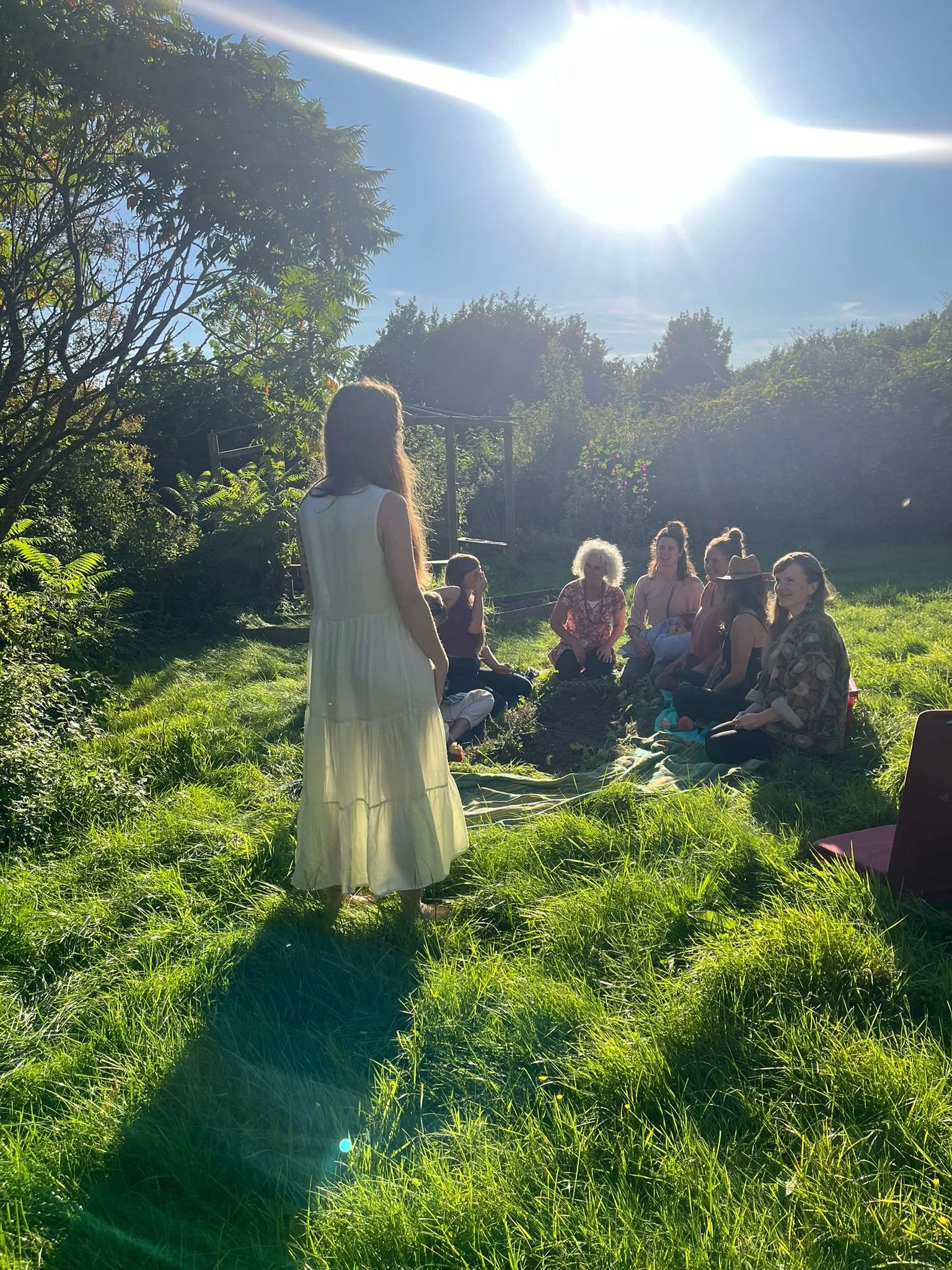 A group of women sitting on the grass in a garden, with one woman standing in front of them. The sun is shining brightly in the sky, creating lens flare and shadows on the grass.