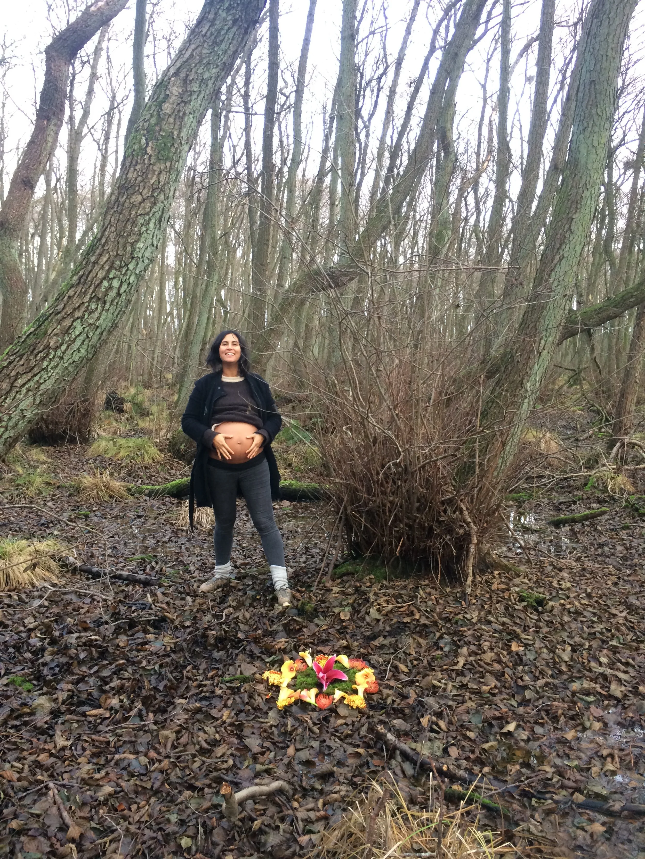 A pregnant woman standing in a wooded area, smiling and holding her belly, with a flower offering on the ground in front of her.