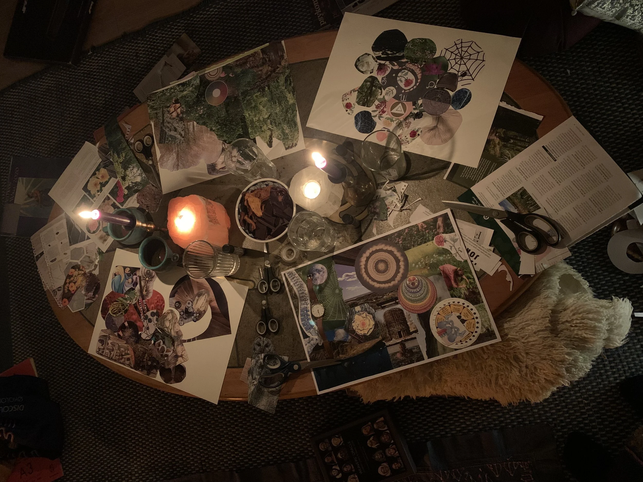 A round wooden table covered with various art projects, pictures, scissors, candles, and glassware. Creating vision boards. 