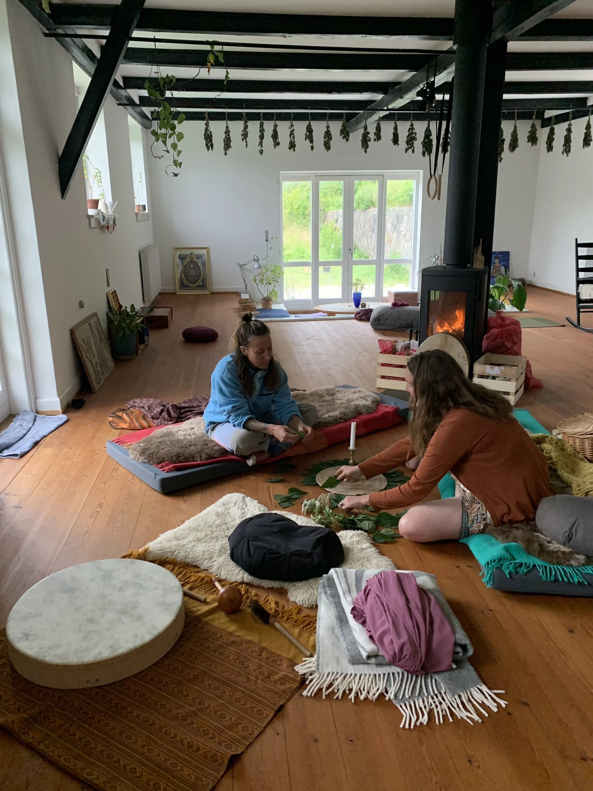 Two women sitting on cushions and blankets on wooden floor, involved in a Stinging Nettle ritual, with a candle, and various objects, in a cozy living room with large windows, a wood stove, and minimal decor.