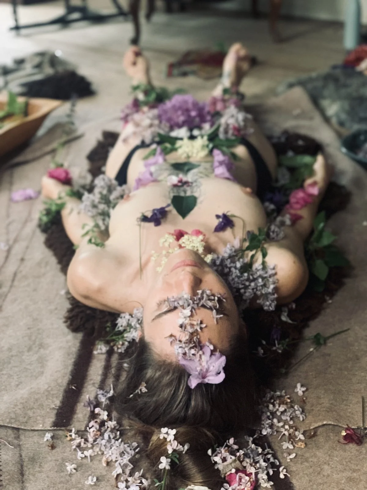 A naked woman lying on the floor covered in colorful flowers and petals, with flowers also in her hair and on her body.